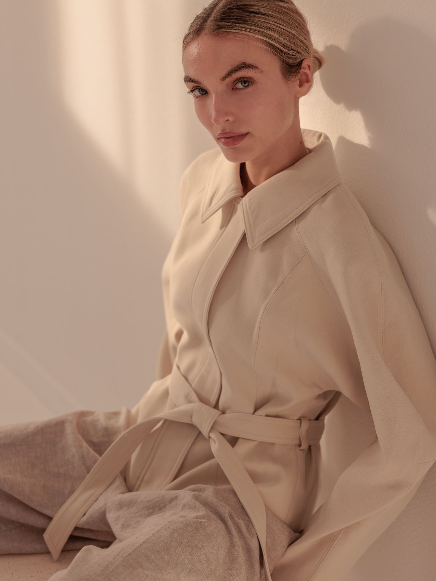 Blonde woman wearing a belted cream coat and grey trousers sitting against a warm sunlit wall.