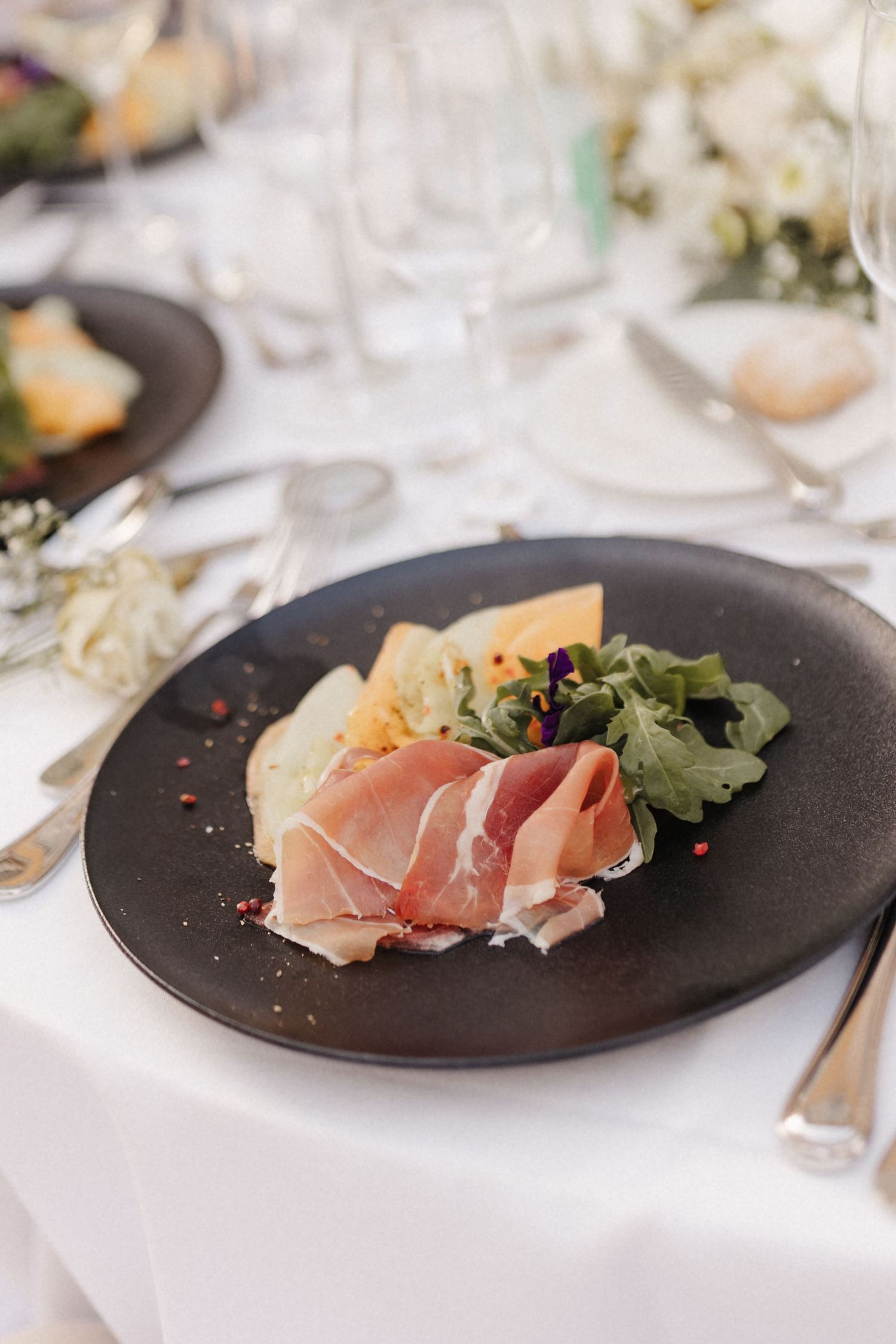 Elegant wedding starter plate with prosciutto, cheese, and greens on a dark ceramic plate with floral table decor.