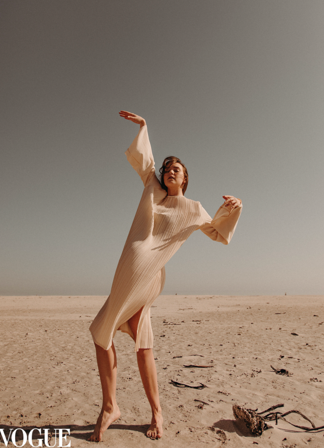 Woman in cream ribbed knit dress posing dynamically on sandy beach under clear sky.
