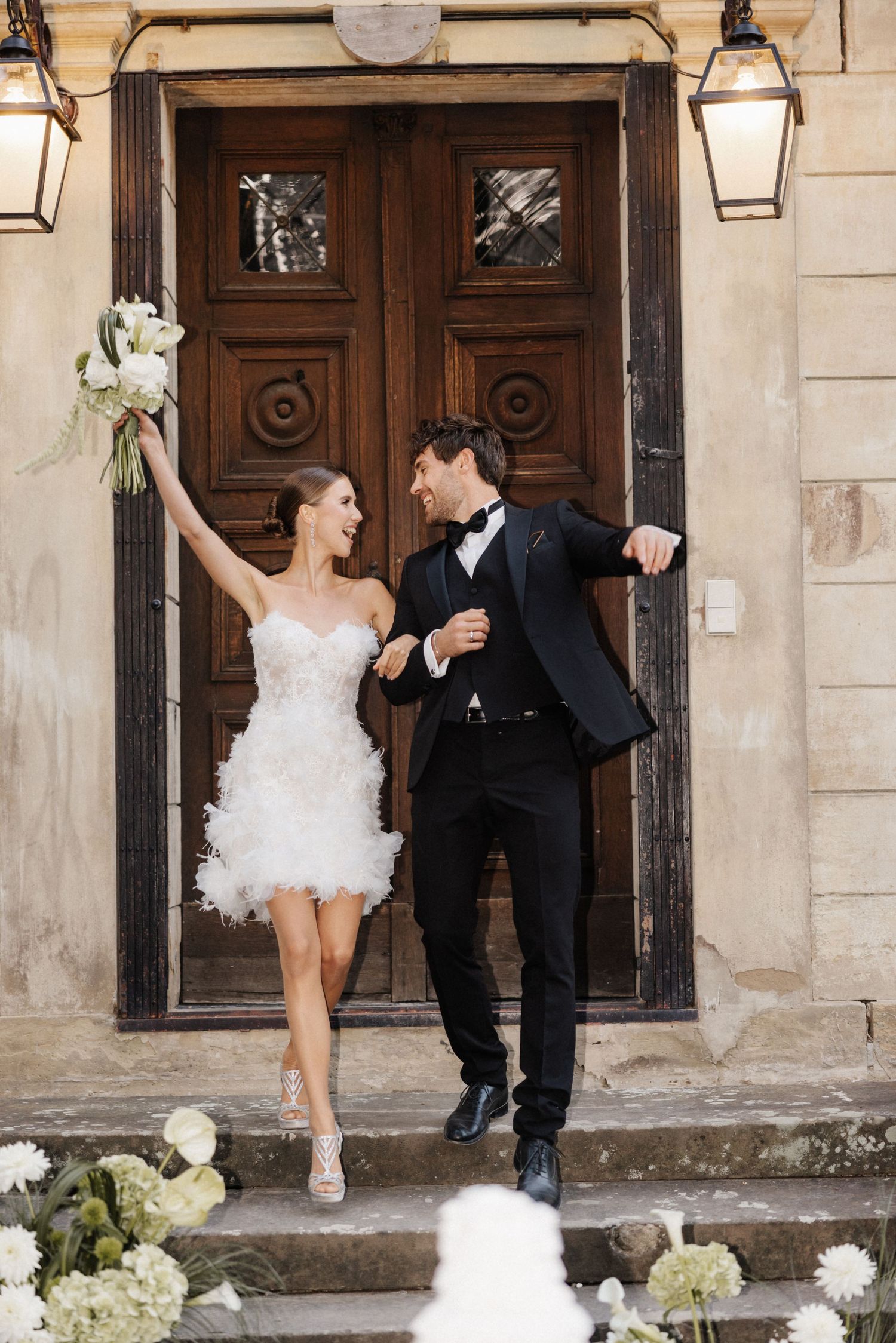 Joyful bride in white feather mini dress and groom in tuxedo celebrating on steps of stone building.