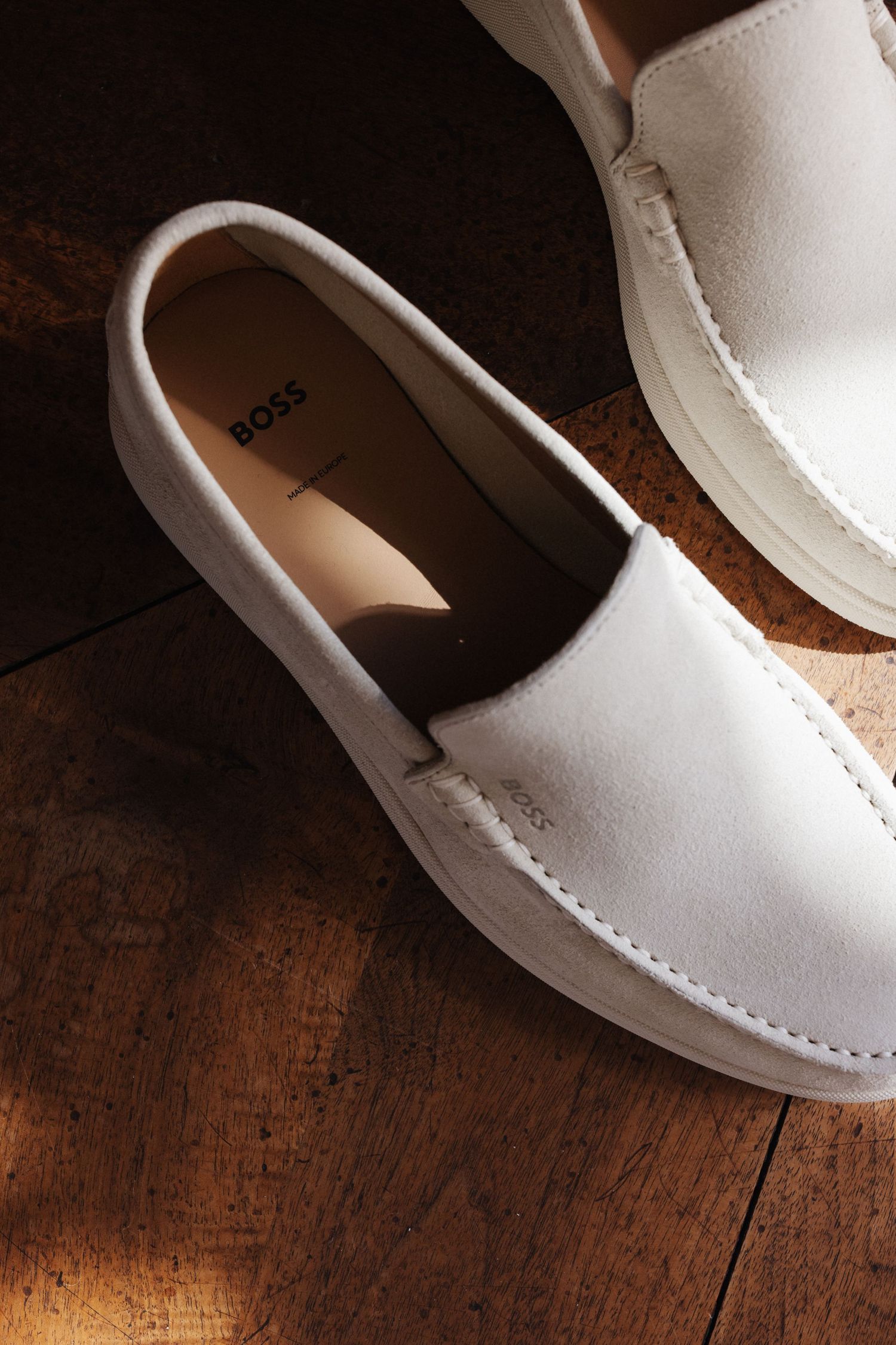 White suede BOSS slip-on loafers on rustic wooden parquet floor in warm natural light.