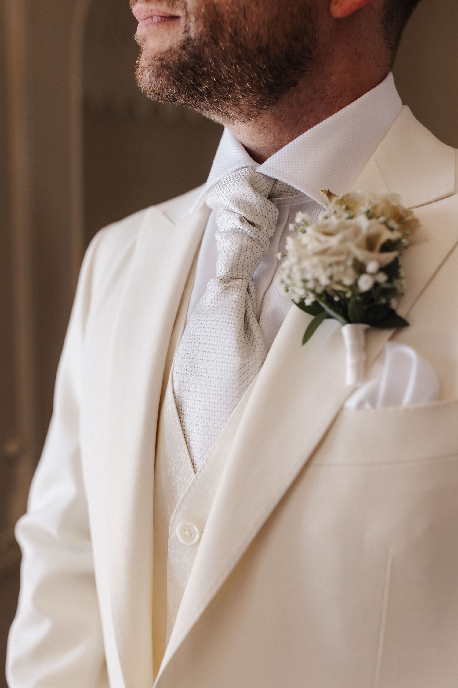 Close-up of groom in cream wedding suit with silver cravat and white floral boutonniere.