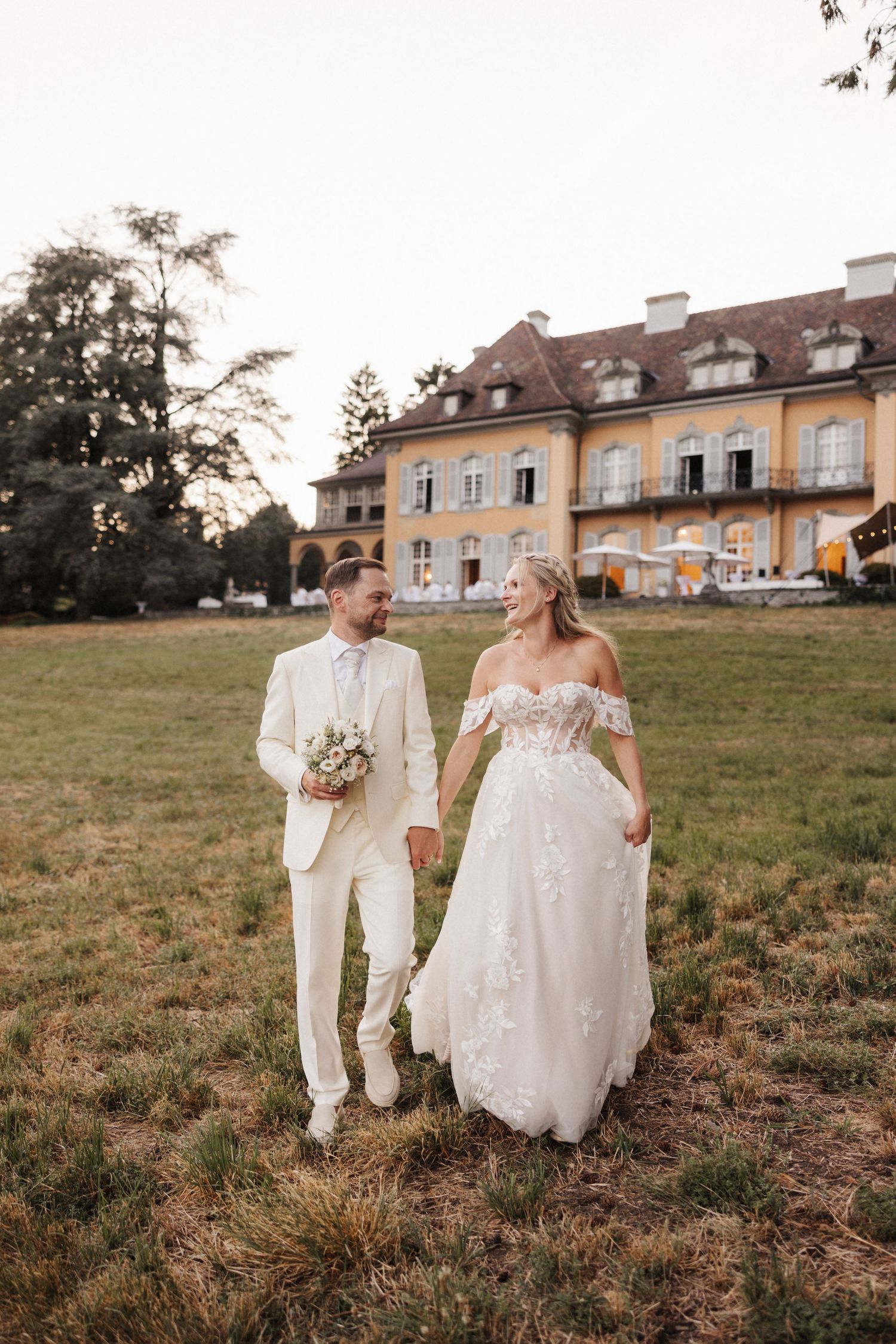 Smiling bride and groom walking together across a grassy field with a yellow European manor house behind them.