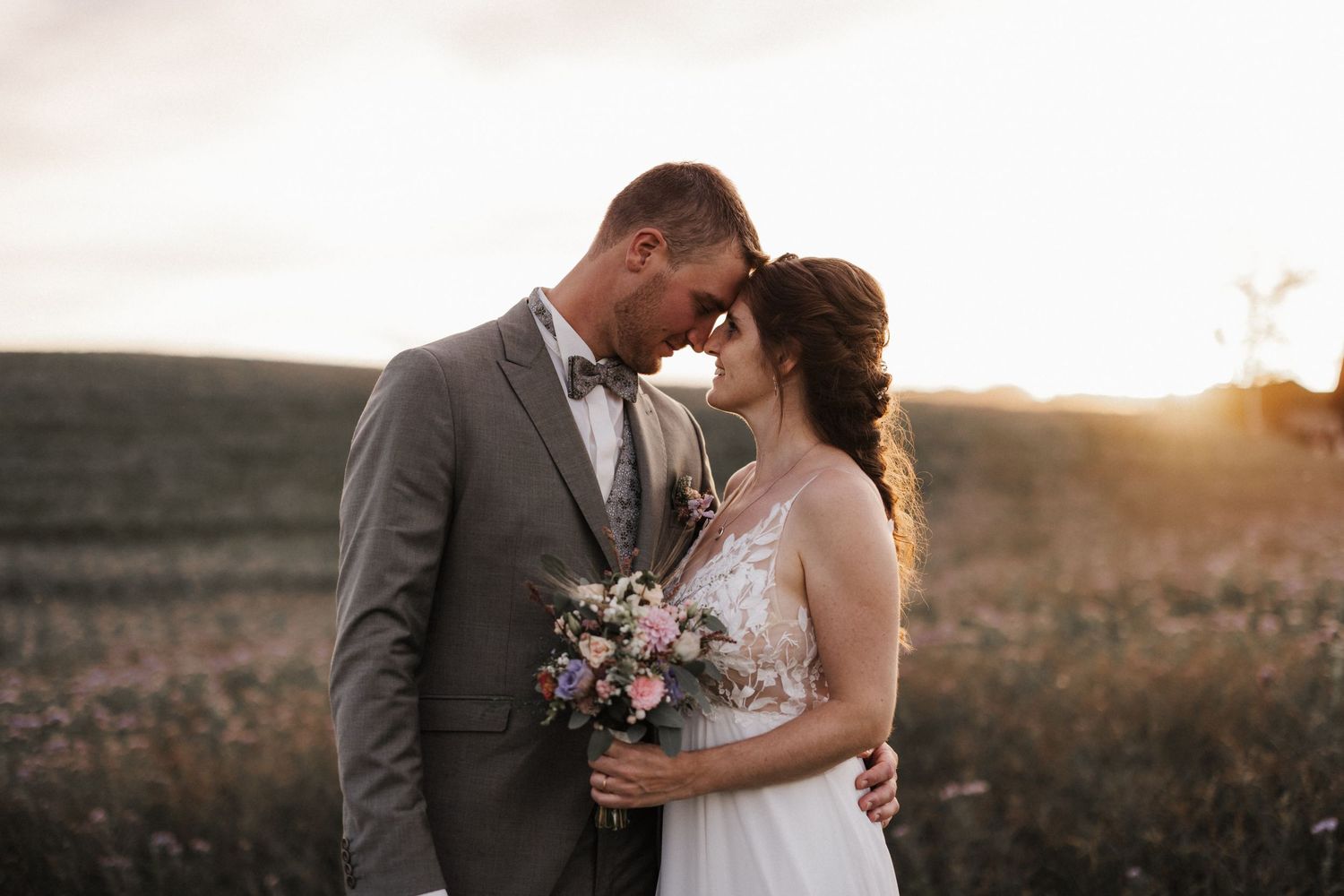 Romantic wedding couple touching foreheads tenderly at golden sunset in an open countryside field.