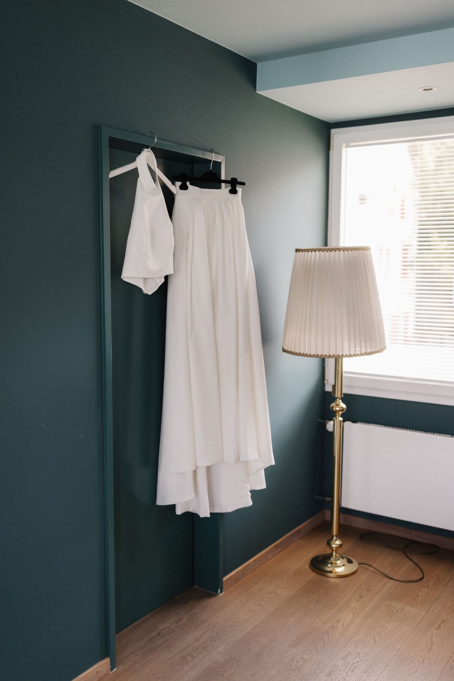 White wedding dress hanging on a rack beside a gold floor lamp in a teal-walled room.
