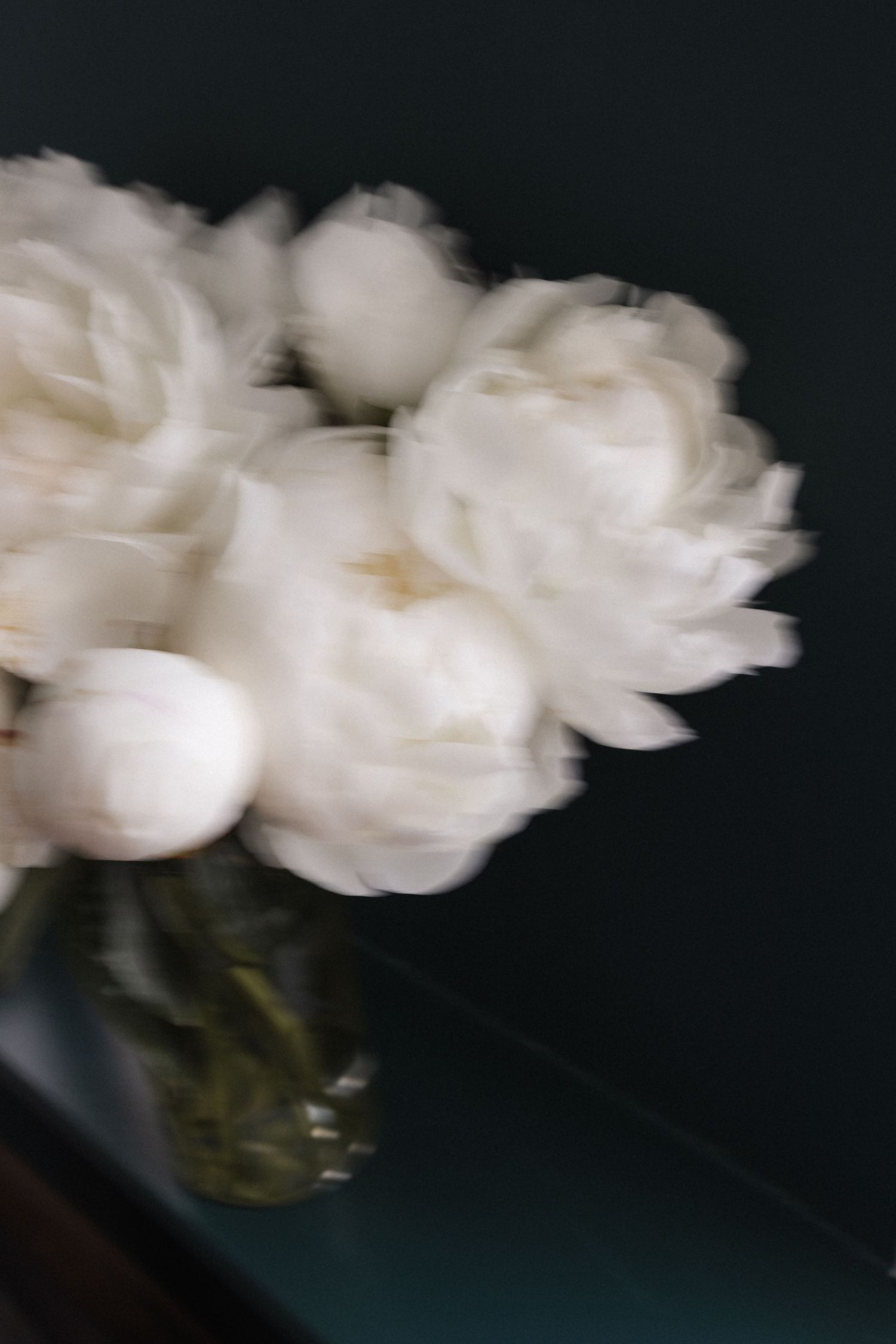 Artistic blurred shot of white peonies in a glass vase on a dark surface.