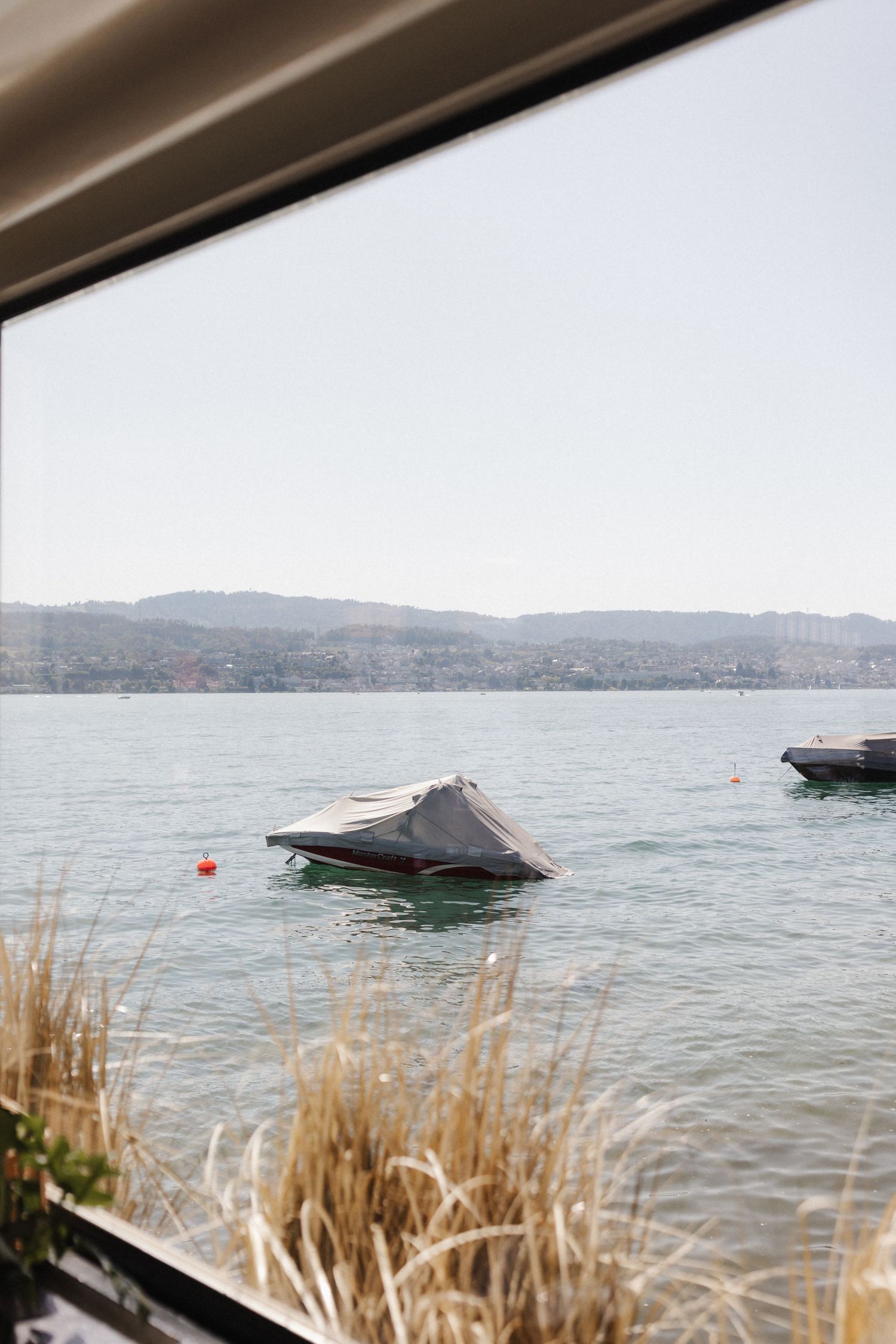 View of calm lake with covered boats and distant mountains through a large open window.