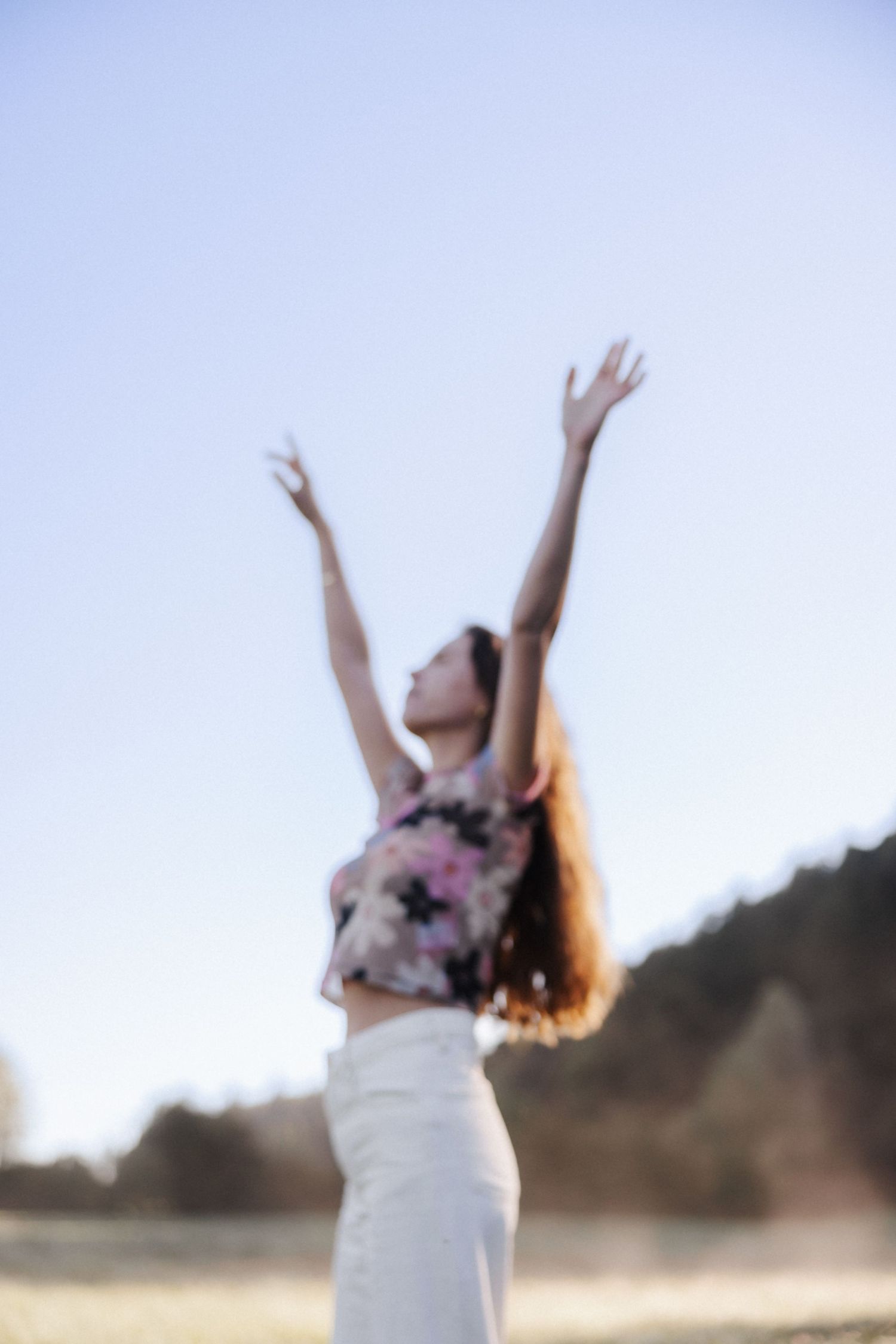 Blurred woman raising arms joyfully toward clear blue sky in an open sunny outdoor field.