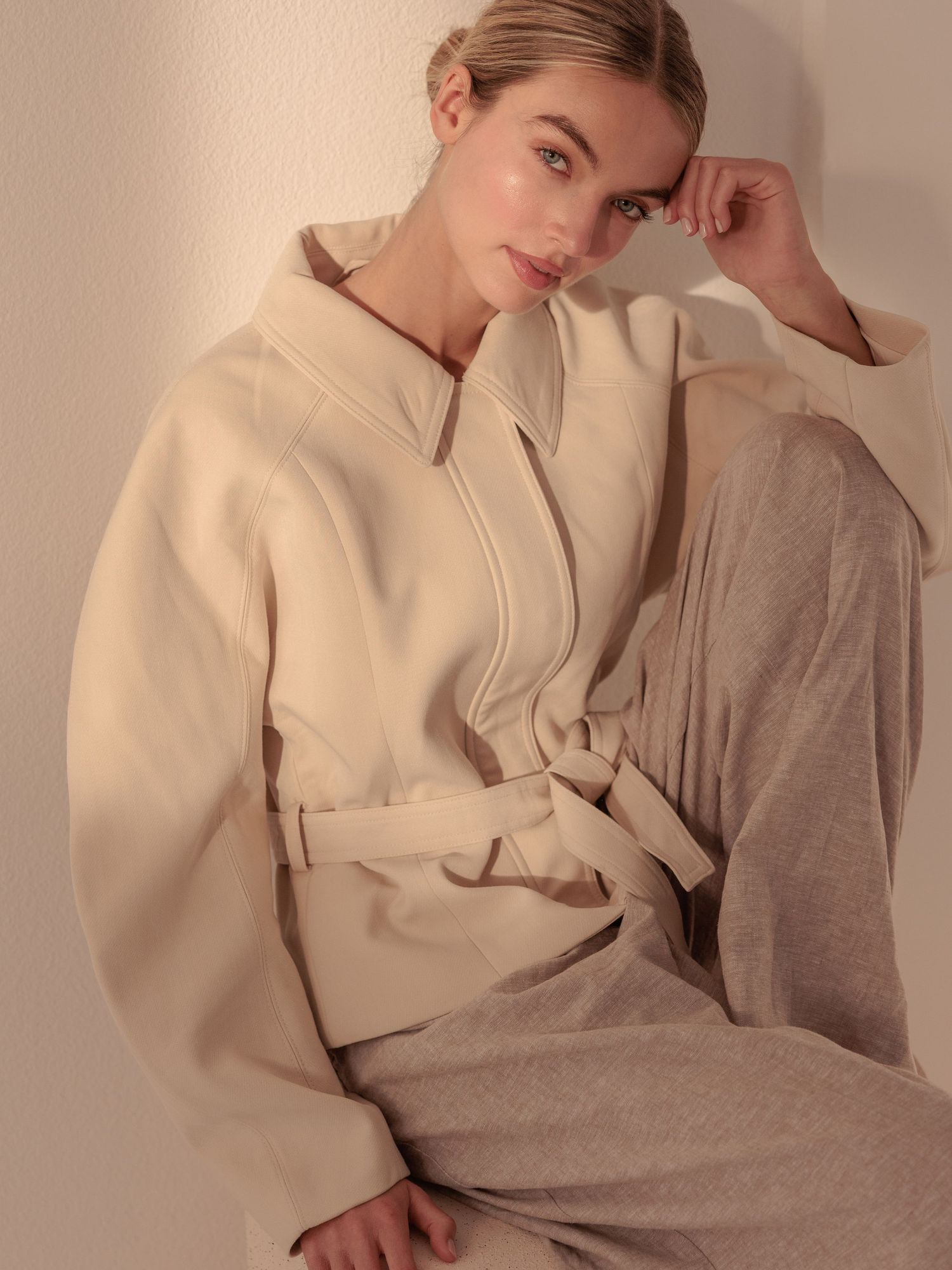 Woman in a cream wrap jacket with belt sits relaxed, resting hand on face in warm light.