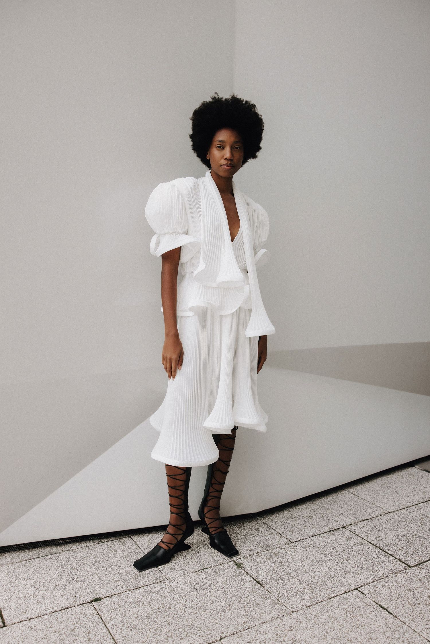 Model wearing a white ruffled midi dress with puff sleeves and black lace-up flat sandals in a minimalist setting.