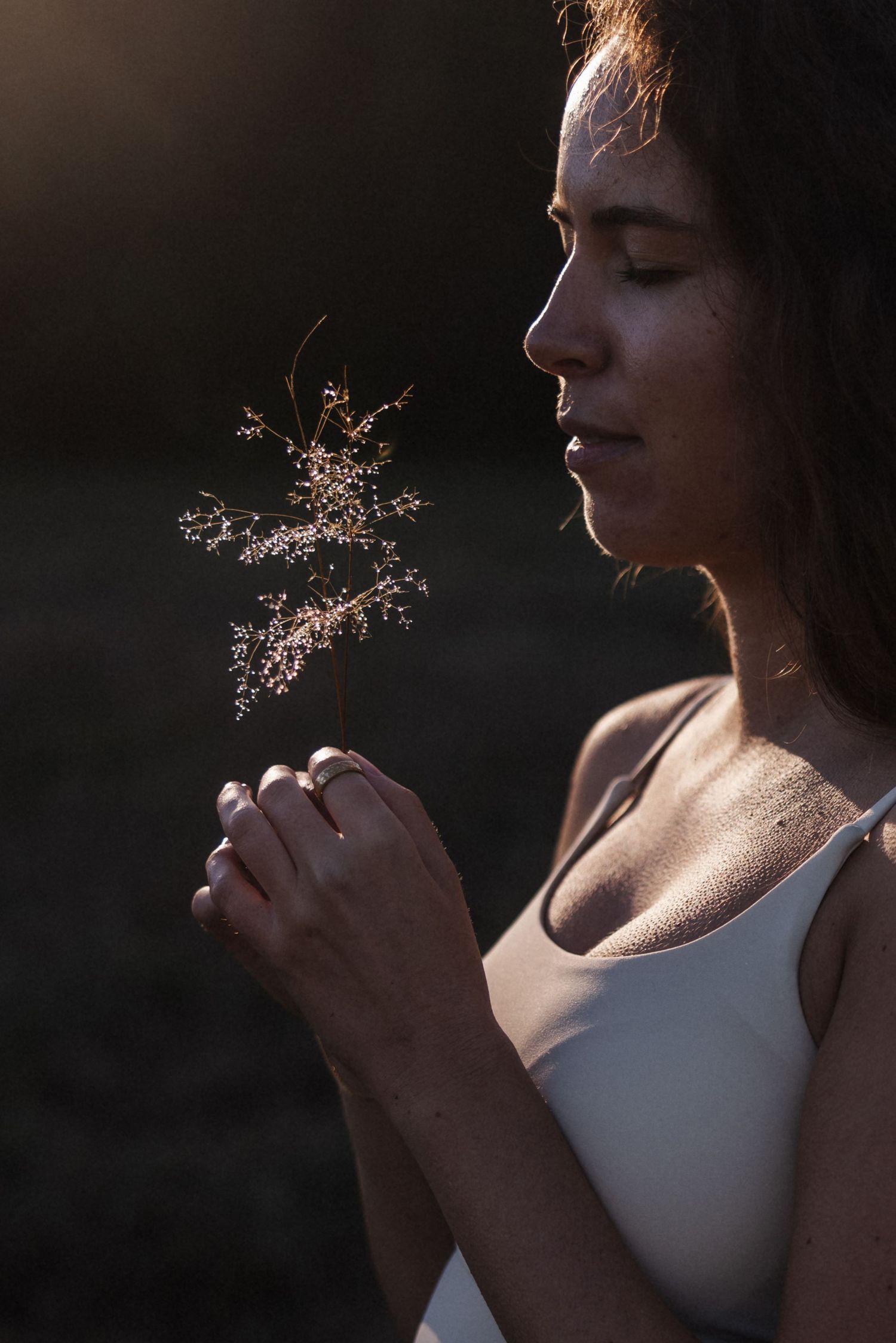 Young woman with closed eyes holds a dried wildflower stem in golden backlit sunlight outdoors.