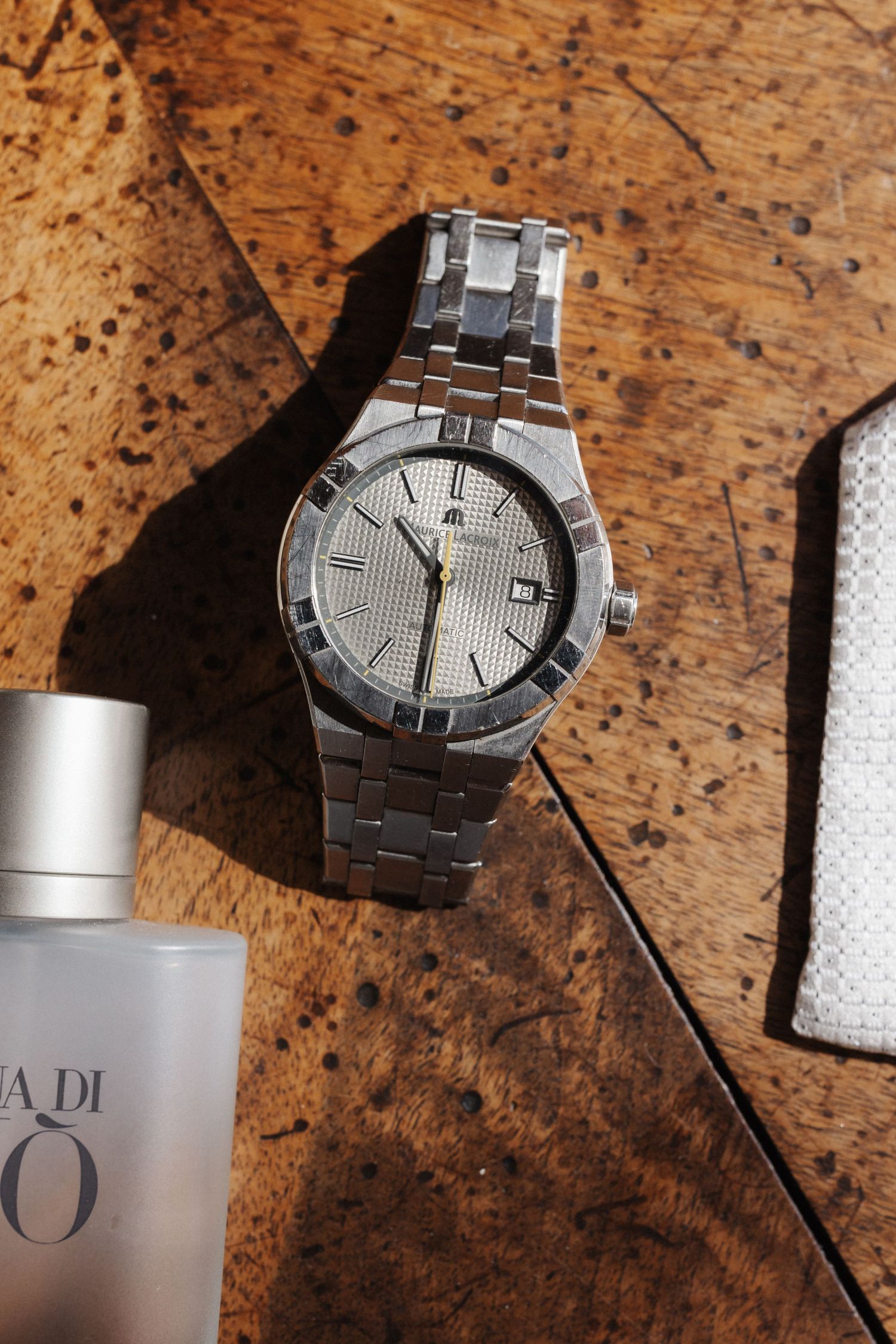 Silver Maurice Lacroix watch on wooden parquet floor beside Acqua di Gio cologne bottle.