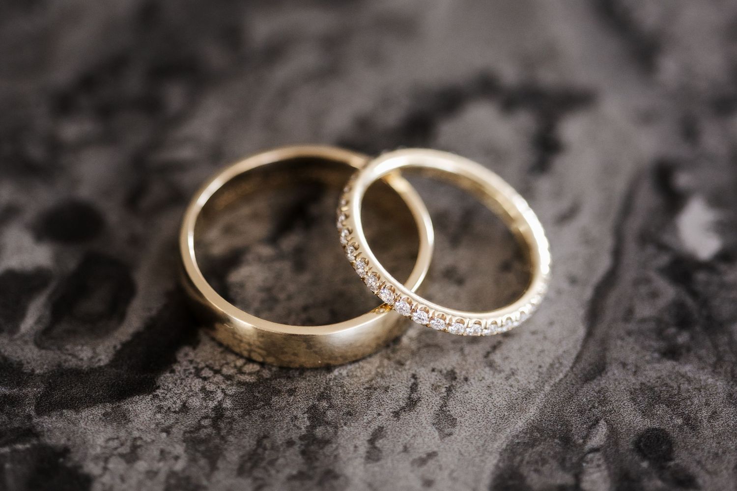 Two gold wedding bands with diamond etching resting on a dark textured stone surface.