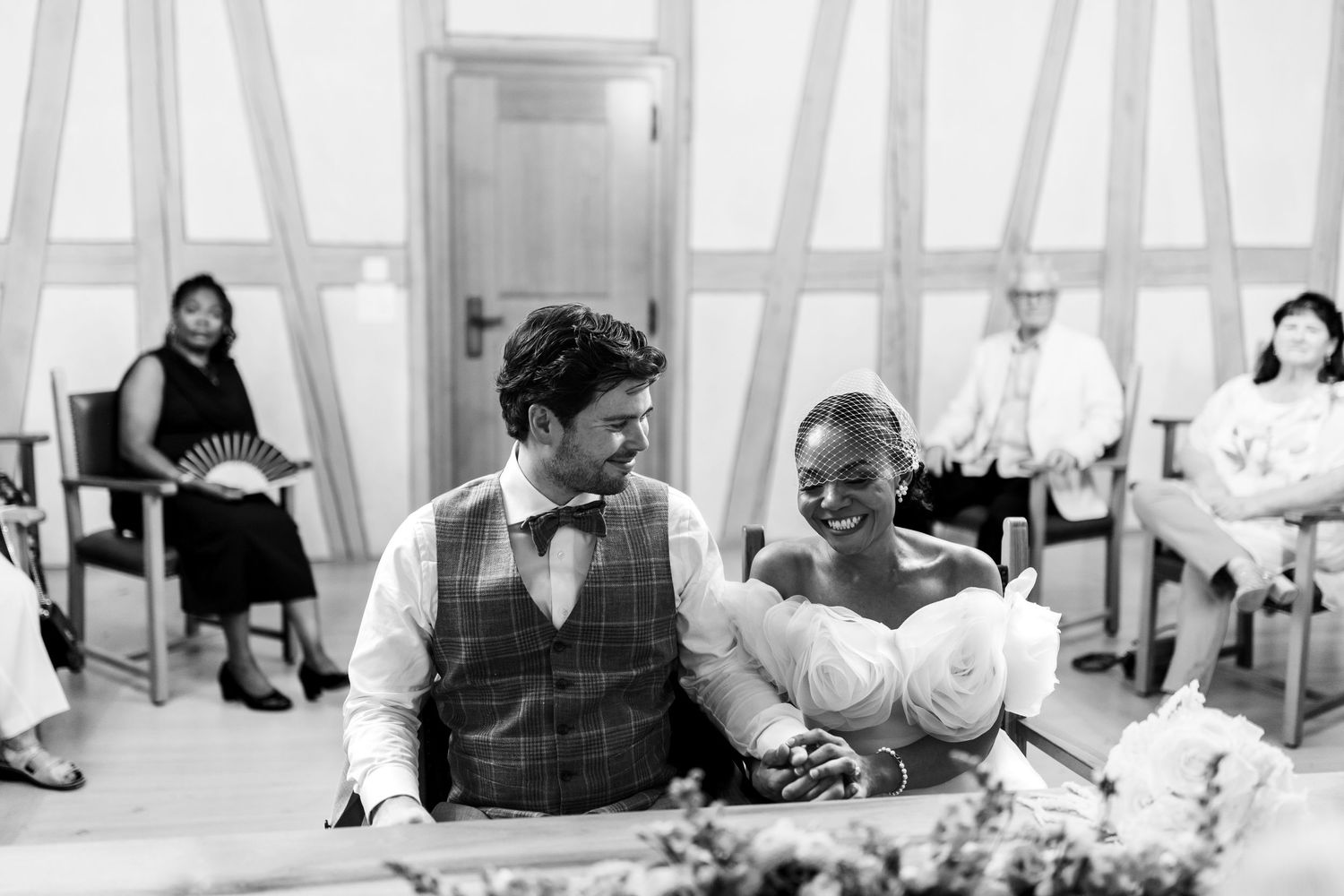 Black and white photo of interracial couple smiling during their wedding ceremony with guests seated.