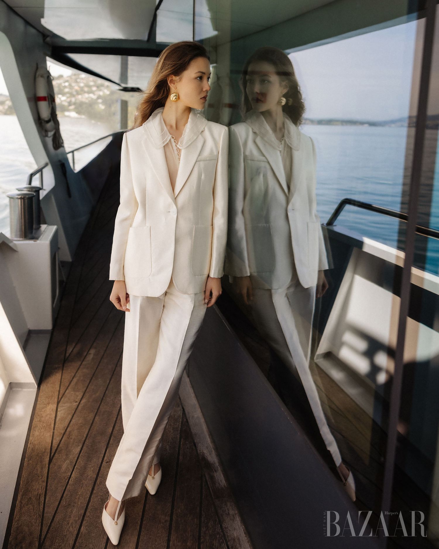 Woman in white blazer suit poses beside glass reflection on a luxury yacht deck.