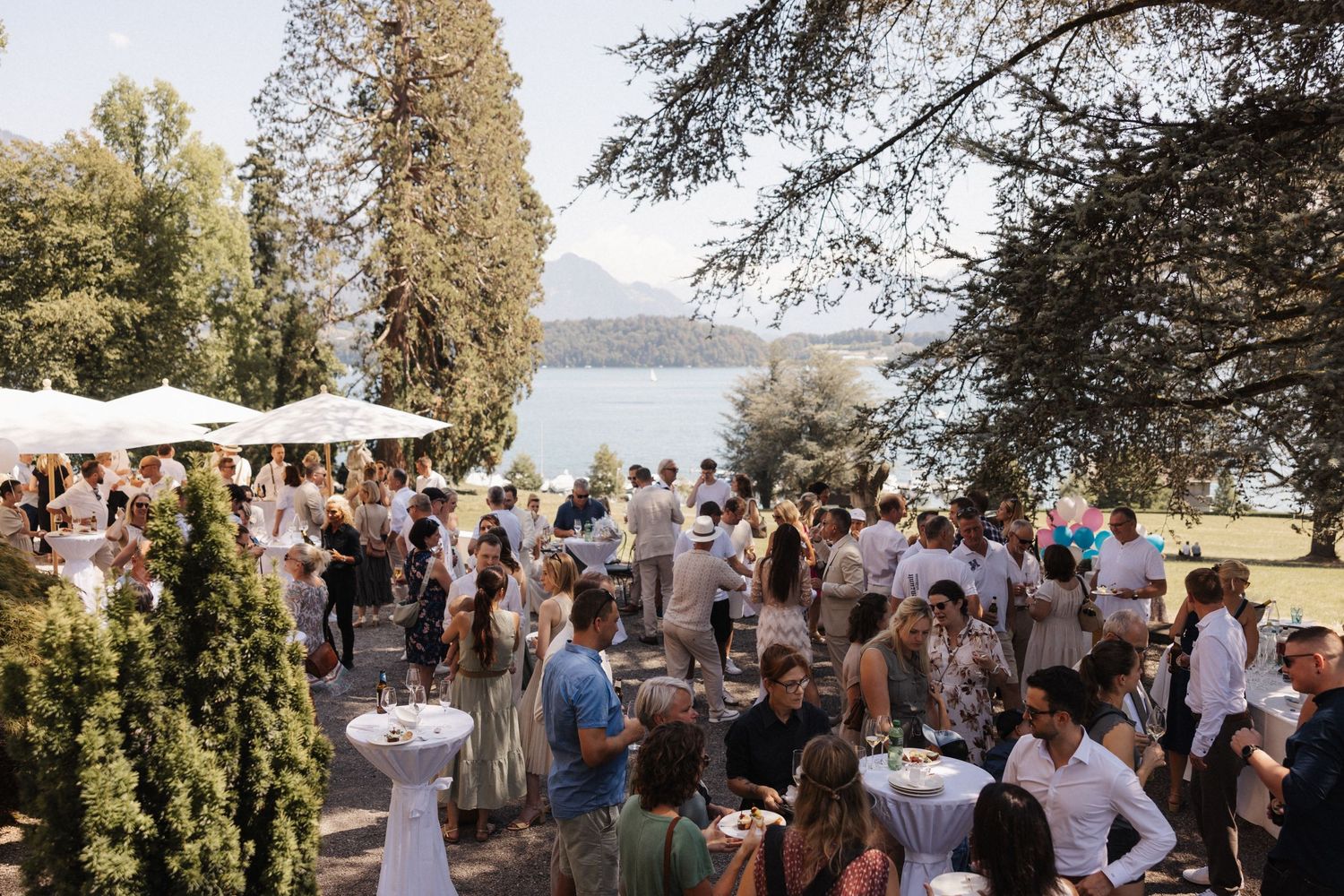 Elegant outdoor wedding reception with guests mingling near a scenic lakeside venue with mountain views.