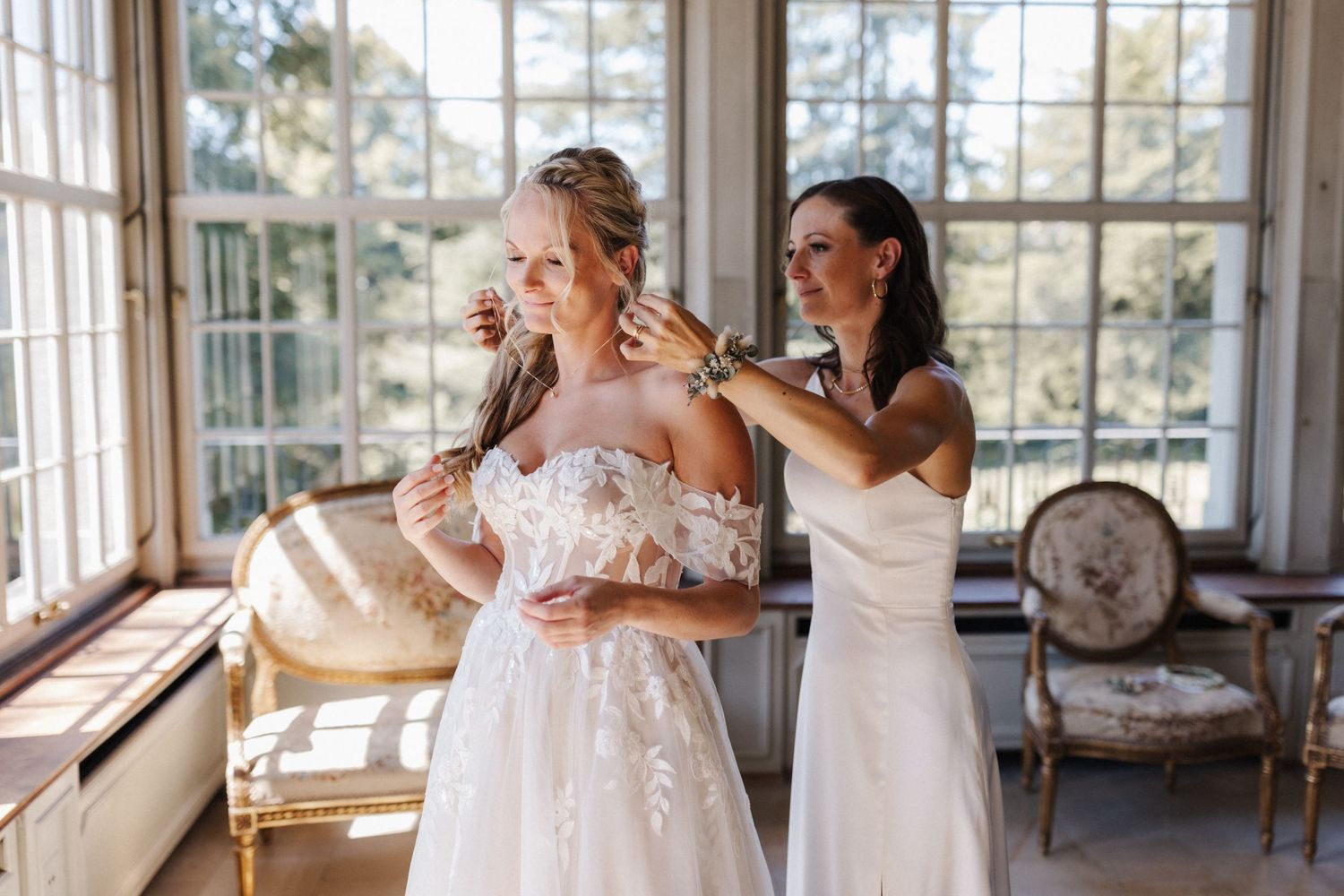 Bridesmaid in white dress helping blonde bride with jewelry in elegant sunlit room with vintage furniture.