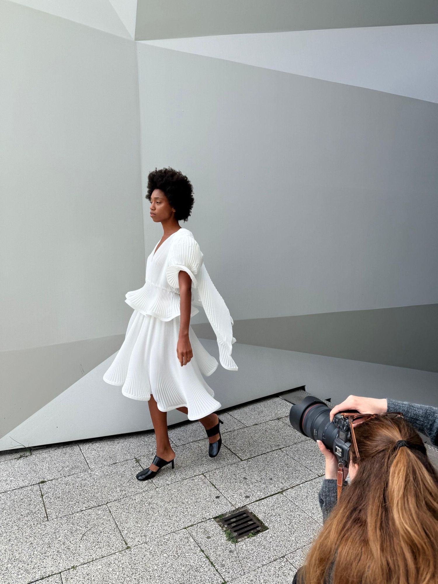 Photographer capturing a model in elegant white ruffled dress outdoors against geometric gray walls.