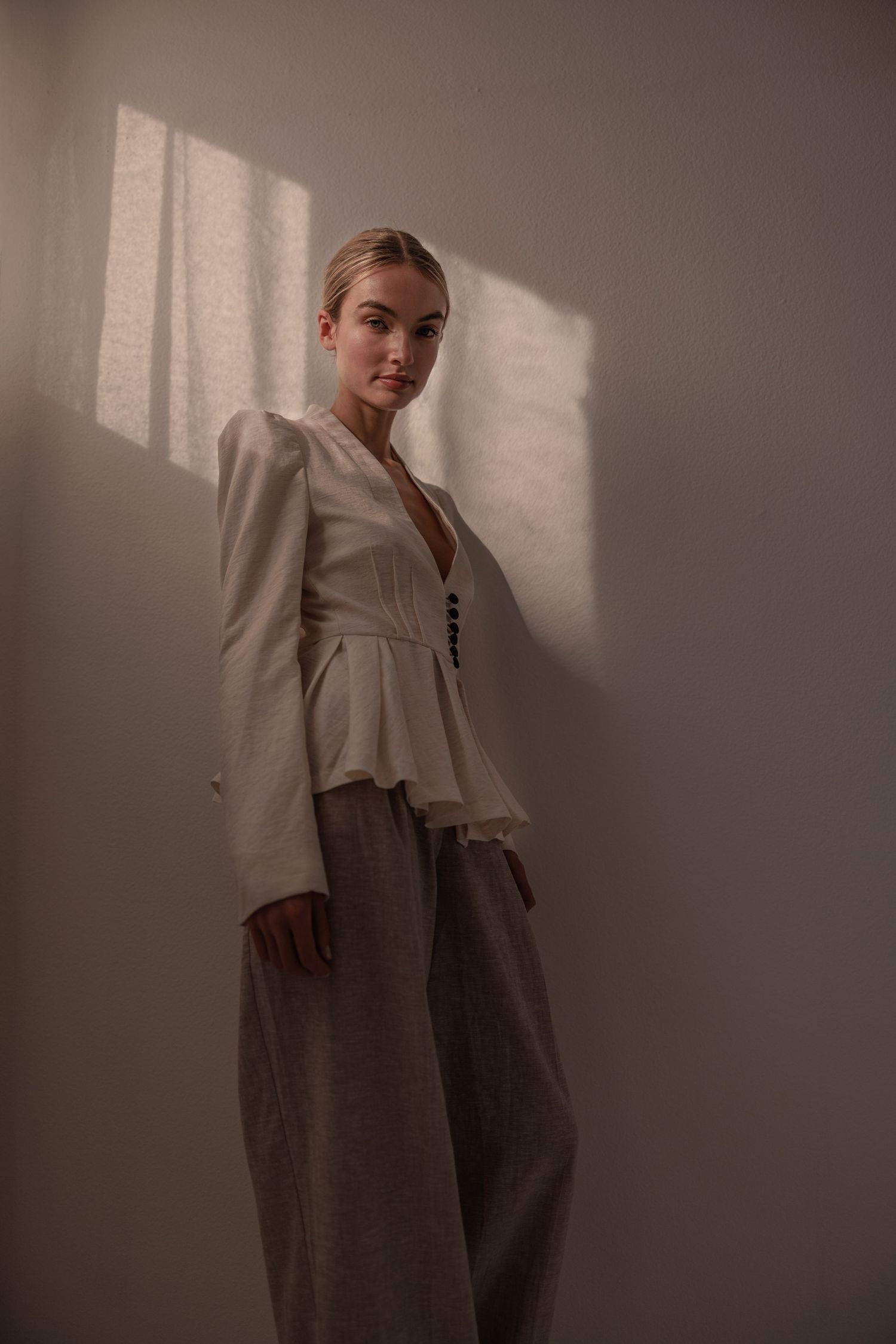 Blonde woman in white peplum blazer and grey wide-leg trousers posing against a sunlit beige wall.