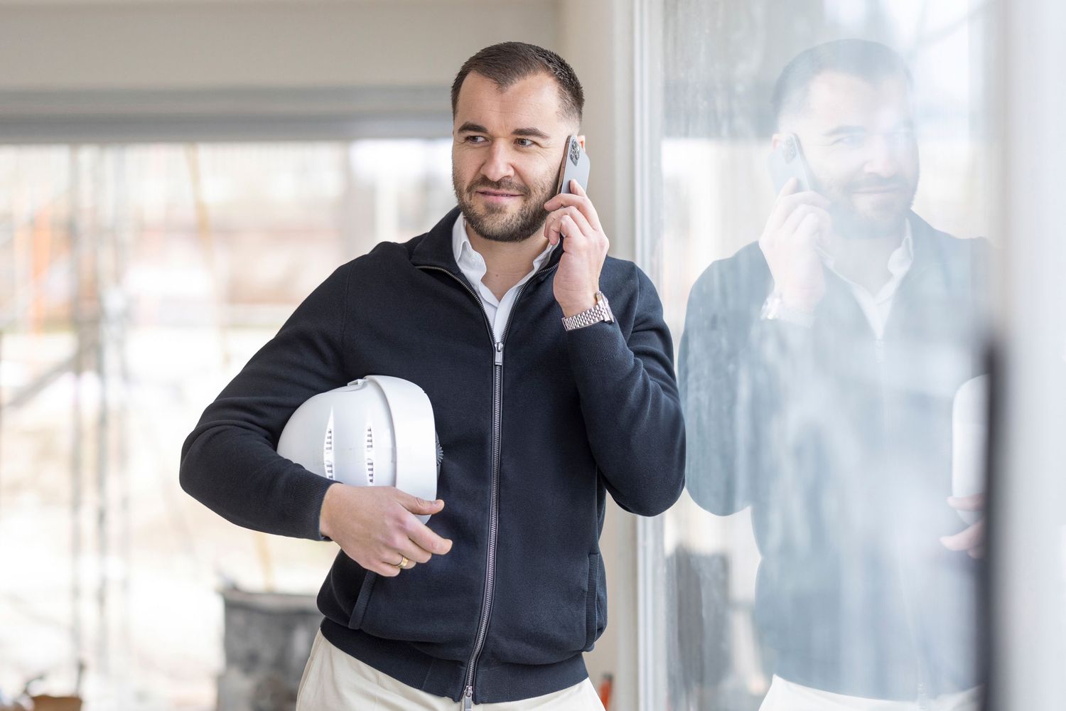Smiling professional in black jacket holding a hard hat while talking on phone near a bright window.