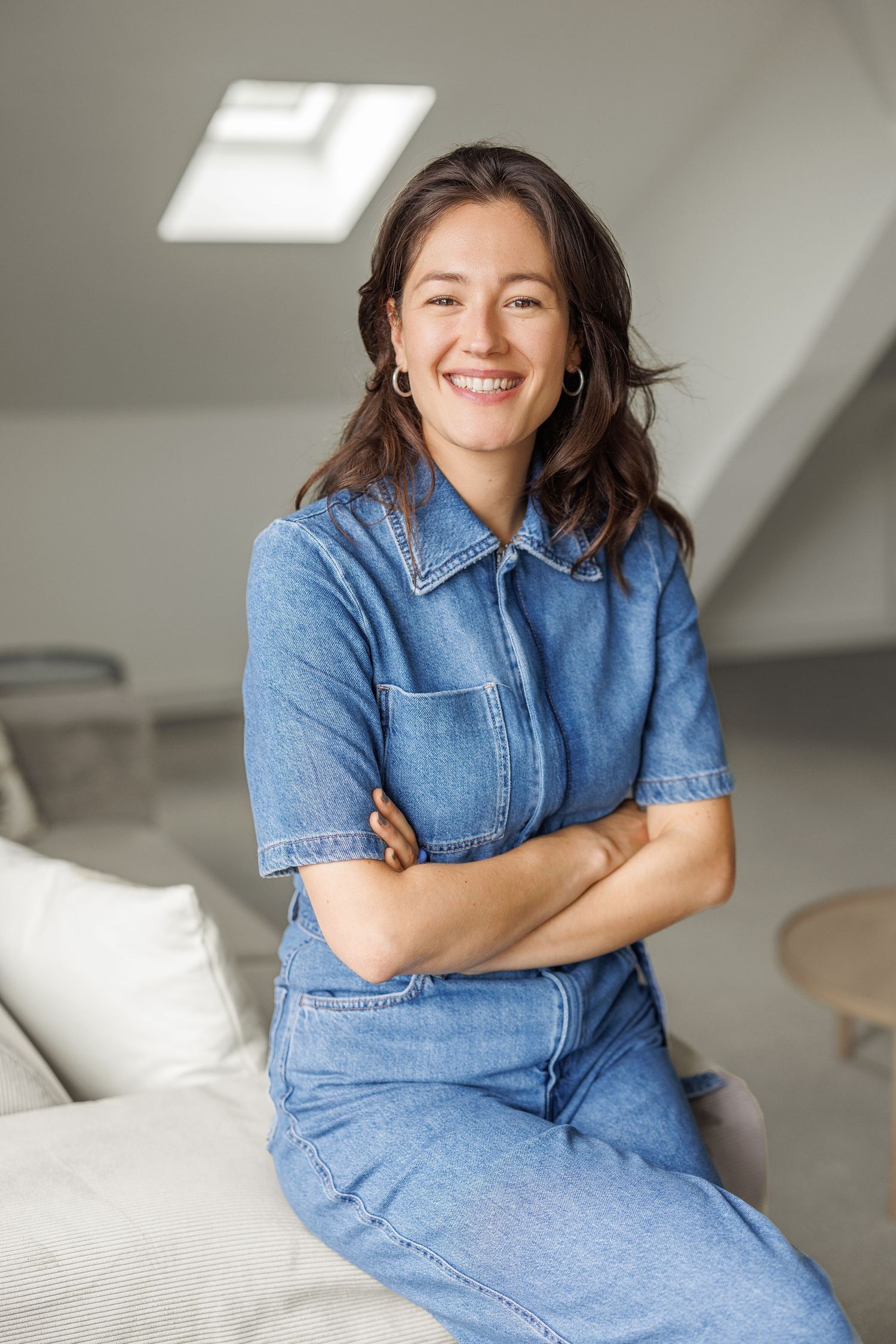 Happy brunette woman in denim jumpsuit sitting on sofa with arms crossed, smiling.