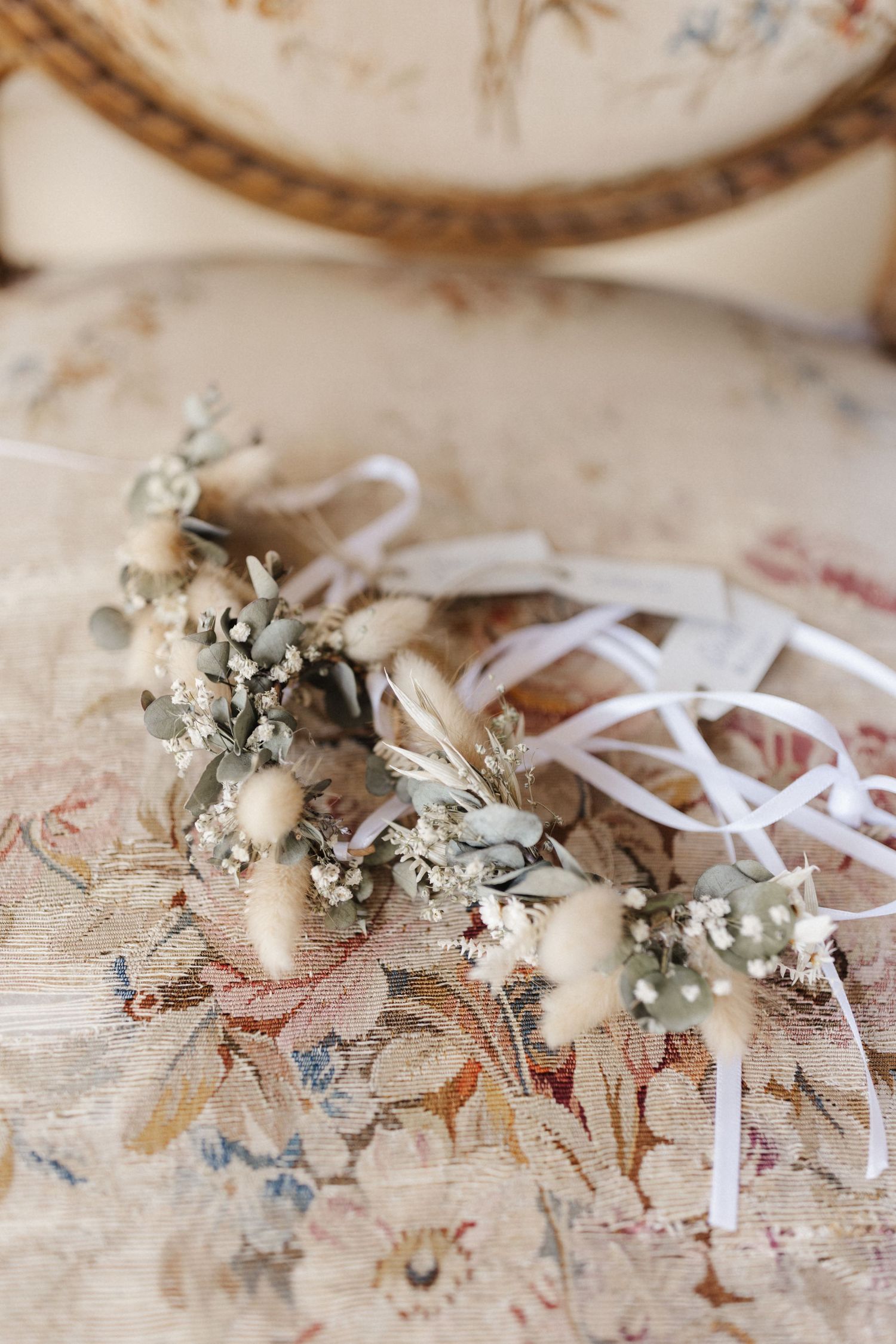 Delicate dried flower crown with eucalyptus, bunny tails, and white ribbon on vintage floral tapestry chair.