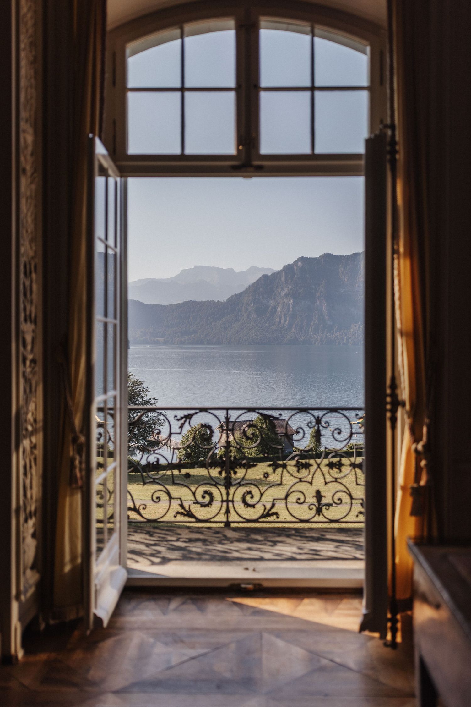 Arched French doors open to ornate iron balcony overlooking serene mountain lake view.