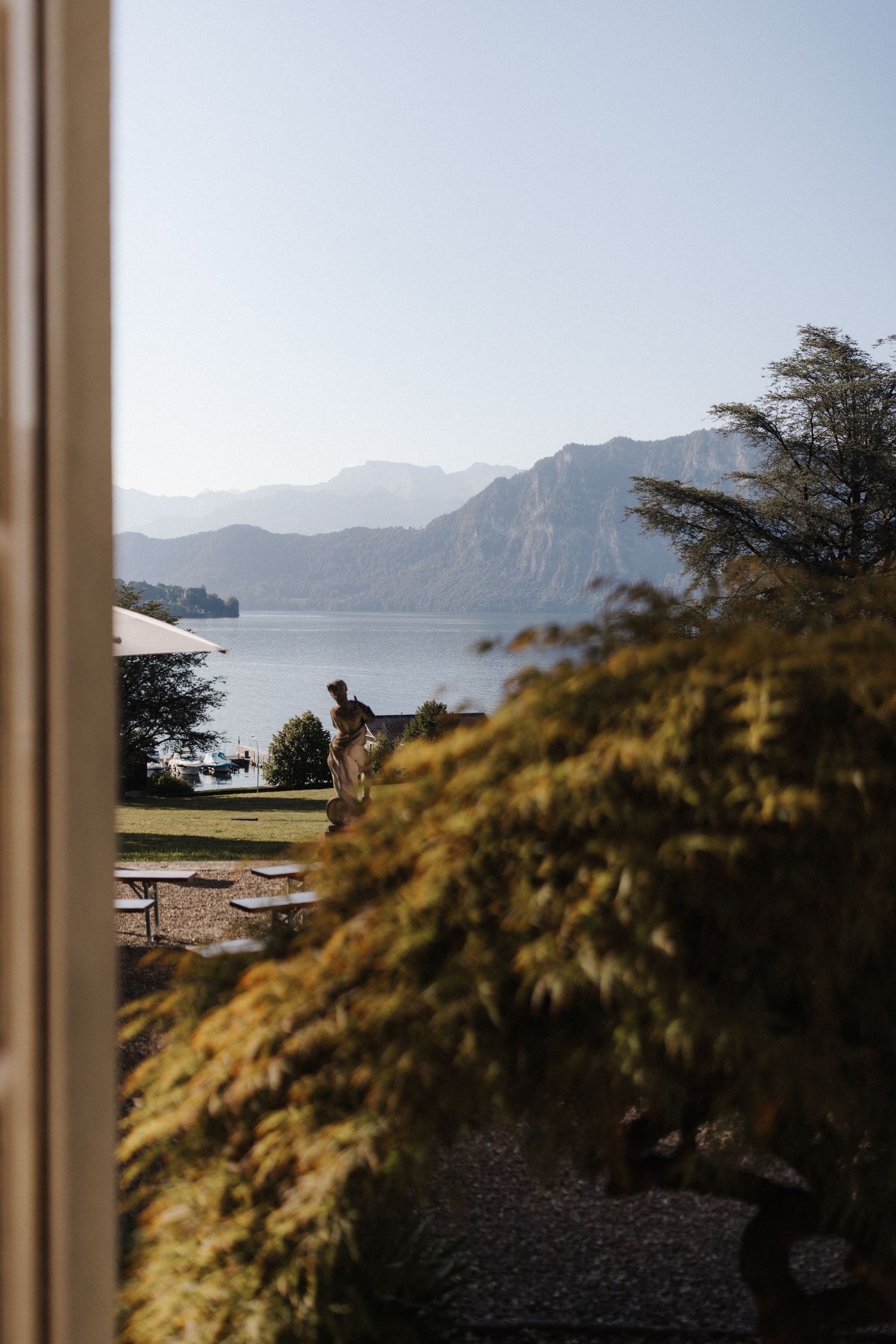 Scenic lakeside view with mountains, statue, and lush greenery under a clear blue sky.