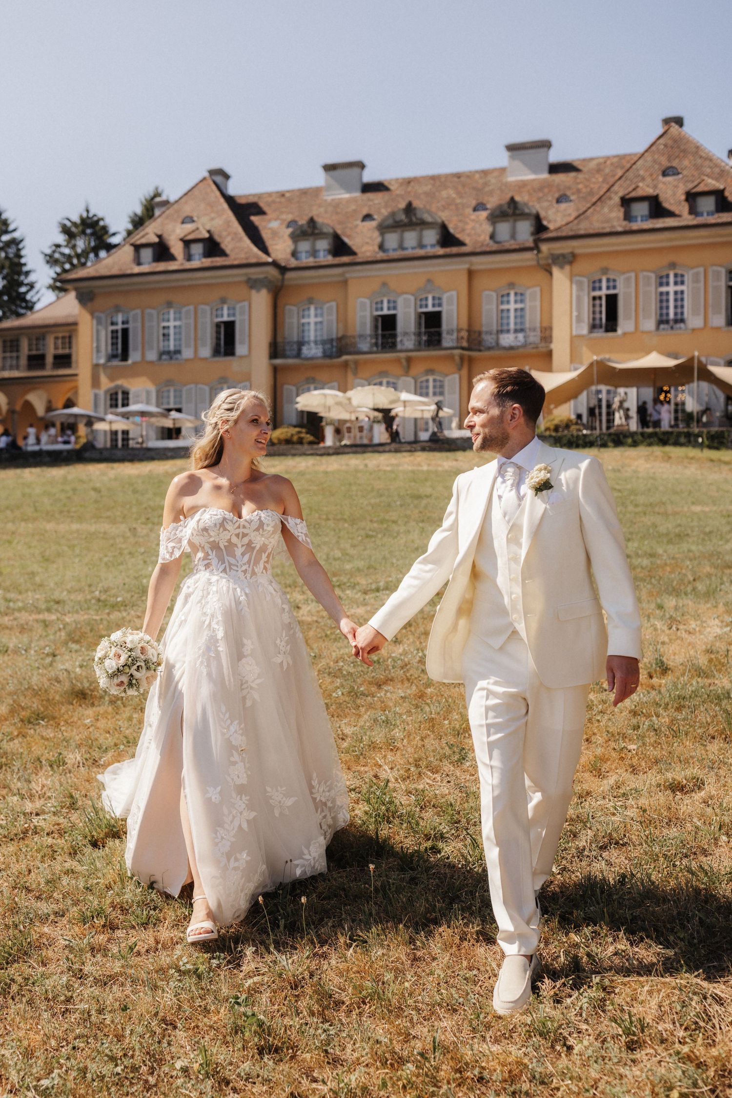 Bride in lace gown and groom in cream suit holding hands walking in front of a grand estate.