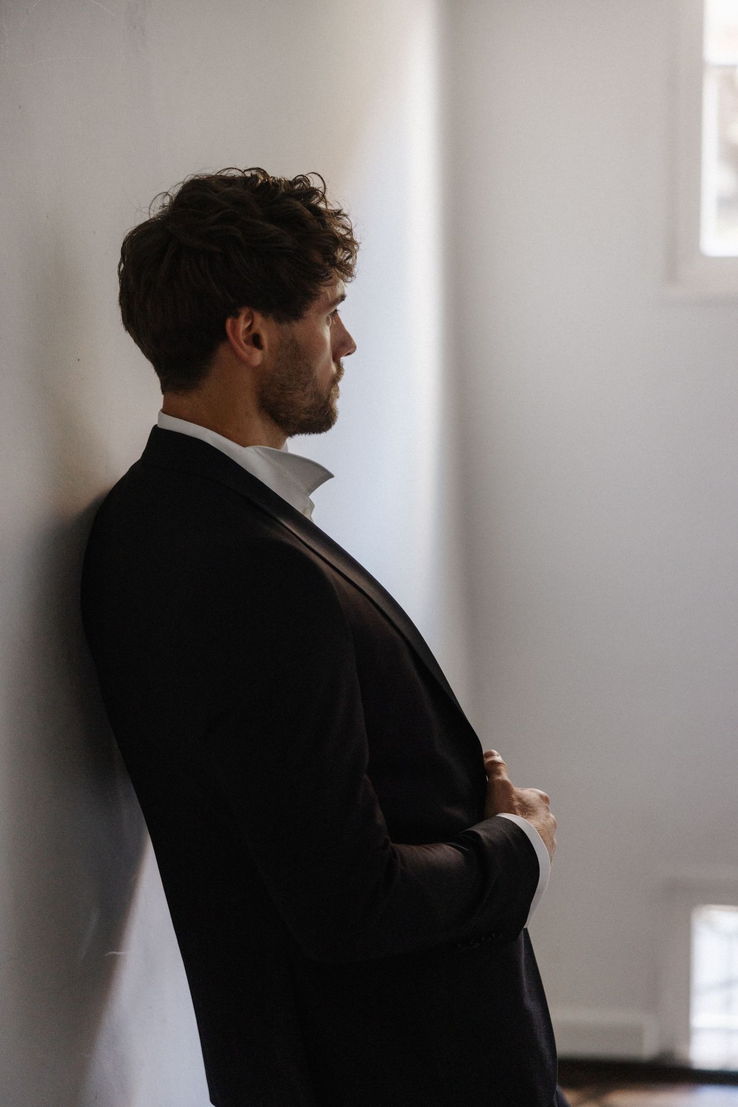 Groom in black suit leaning against white wall with arms crossed in contemplative pose.