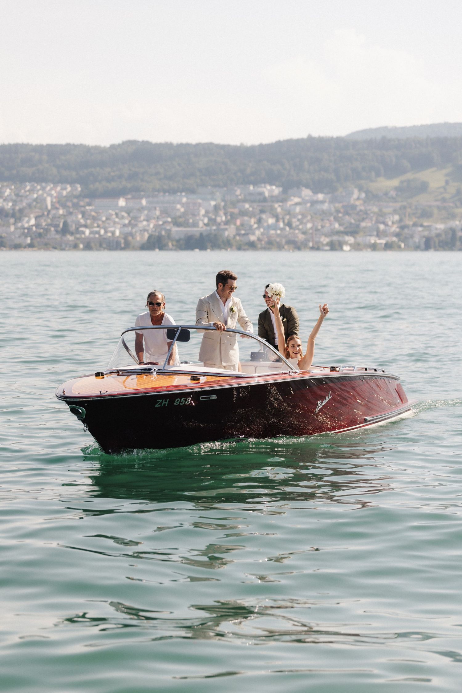 Wedding party celebrating on a vintage wooden motorboat on a turquoise alpine lake with mountains.