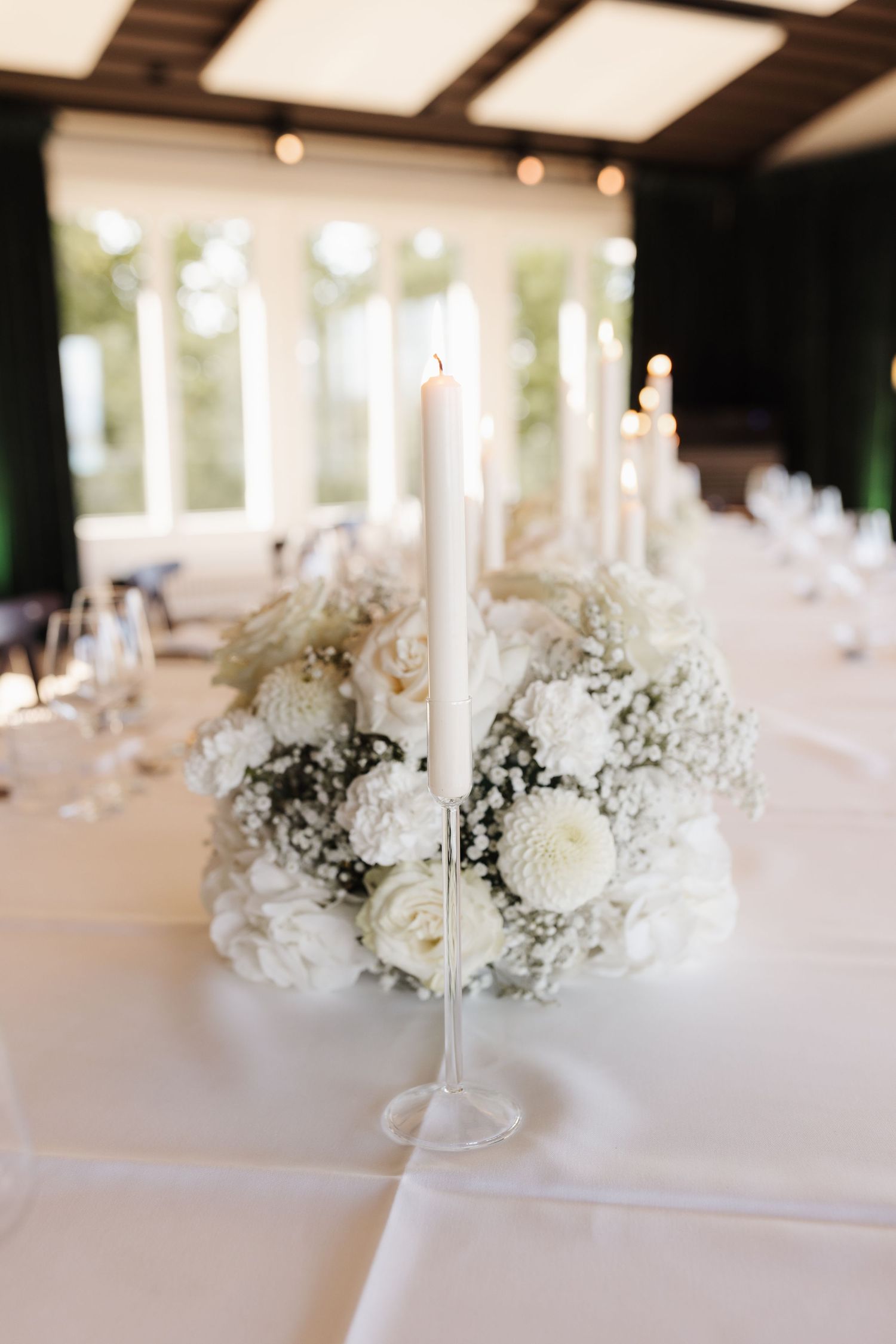 White wedding centerpiece featuring roses, dahlias, and baby's breath surrounding a tall taper candle on a glass holder.