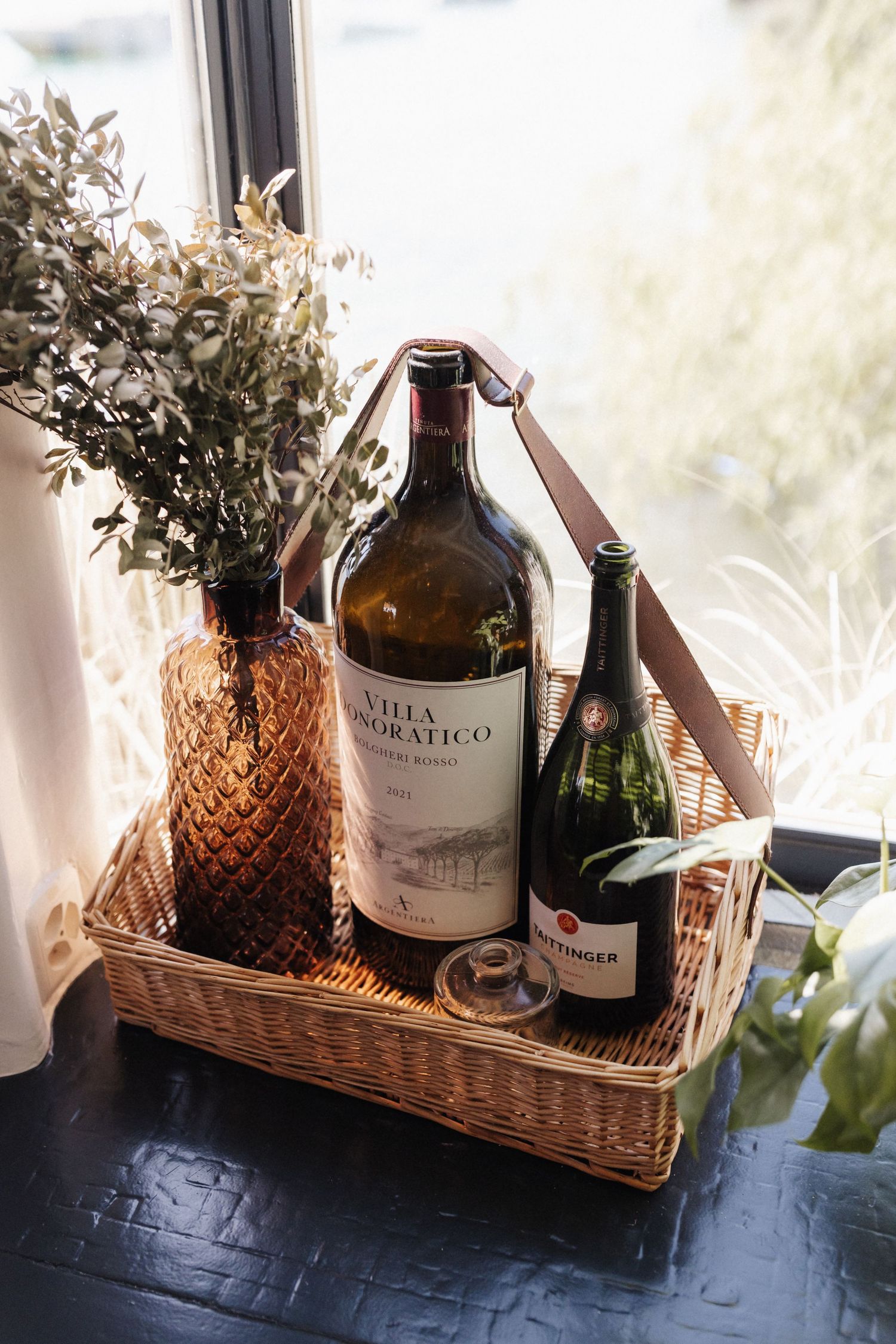 Villa Honoratico wine bottle in a wicker tray with champagne and dried botanics by a window.