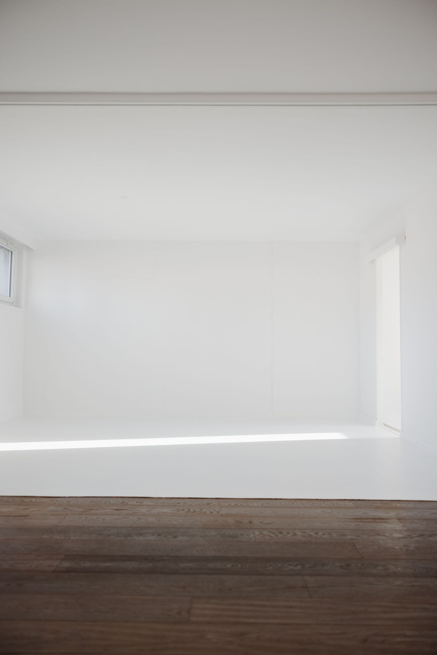 Minimalist white interior room with dark hardwood floors and soft natural light from a low window.