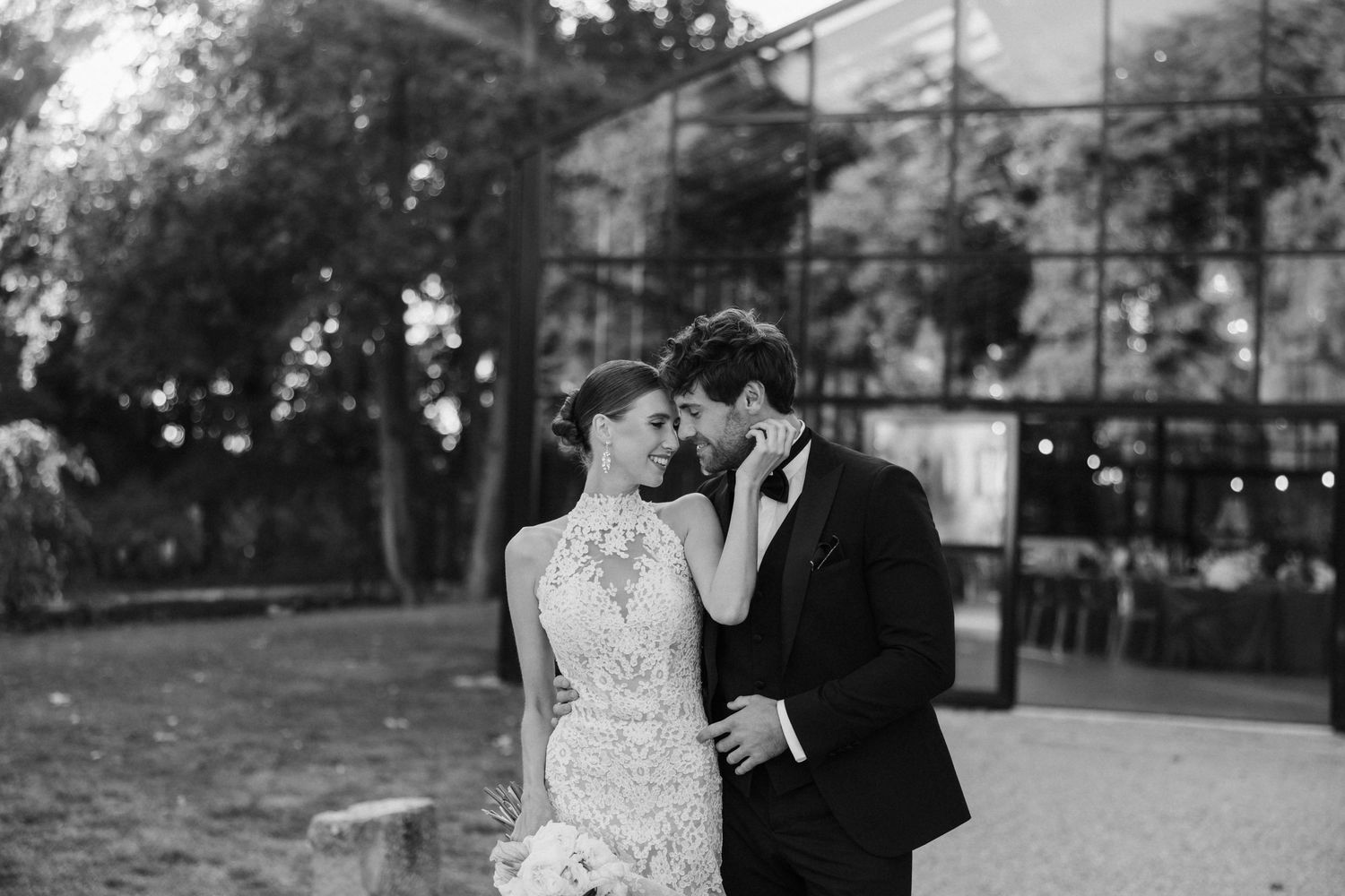 Black and white romantic wedding photo of bride and groom almost kissing outdoors near glass venue.