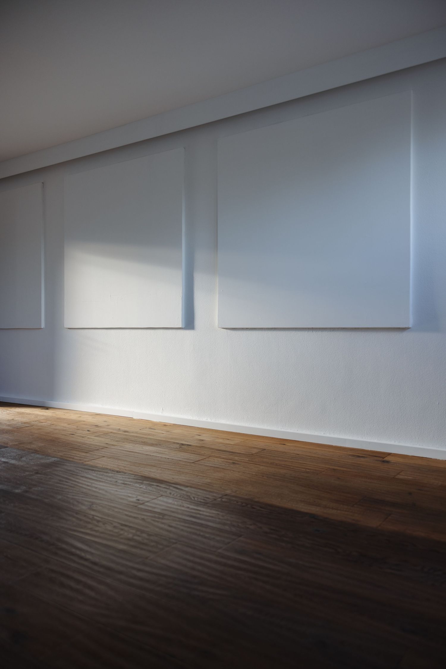 Minimalist white gallery room with two large blank canvases on wall, illuminated by natural sunlight on dark wood floor.