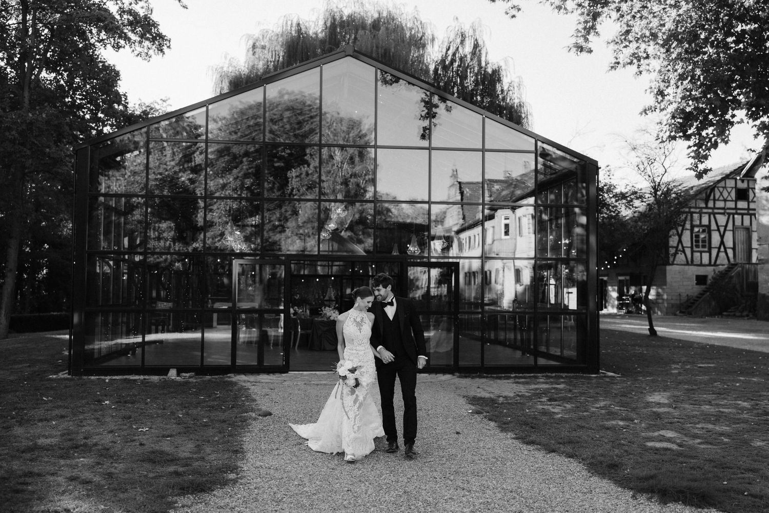 Black and white photo of bride and groom posing in front of a large glass greenhouse structure.