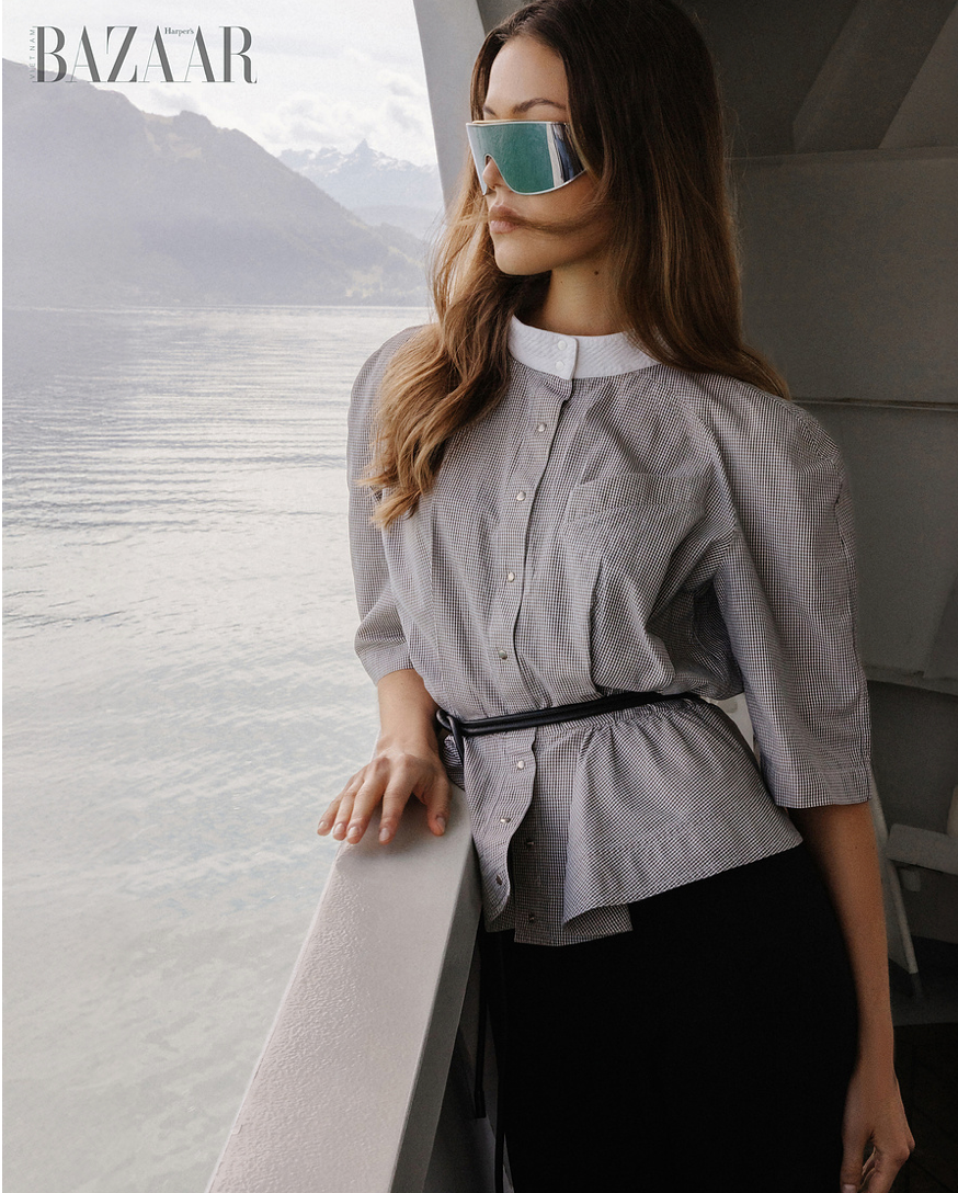 Woman in grey belted peplum shirt and futuristic sunglasses poses on a boat balcony.