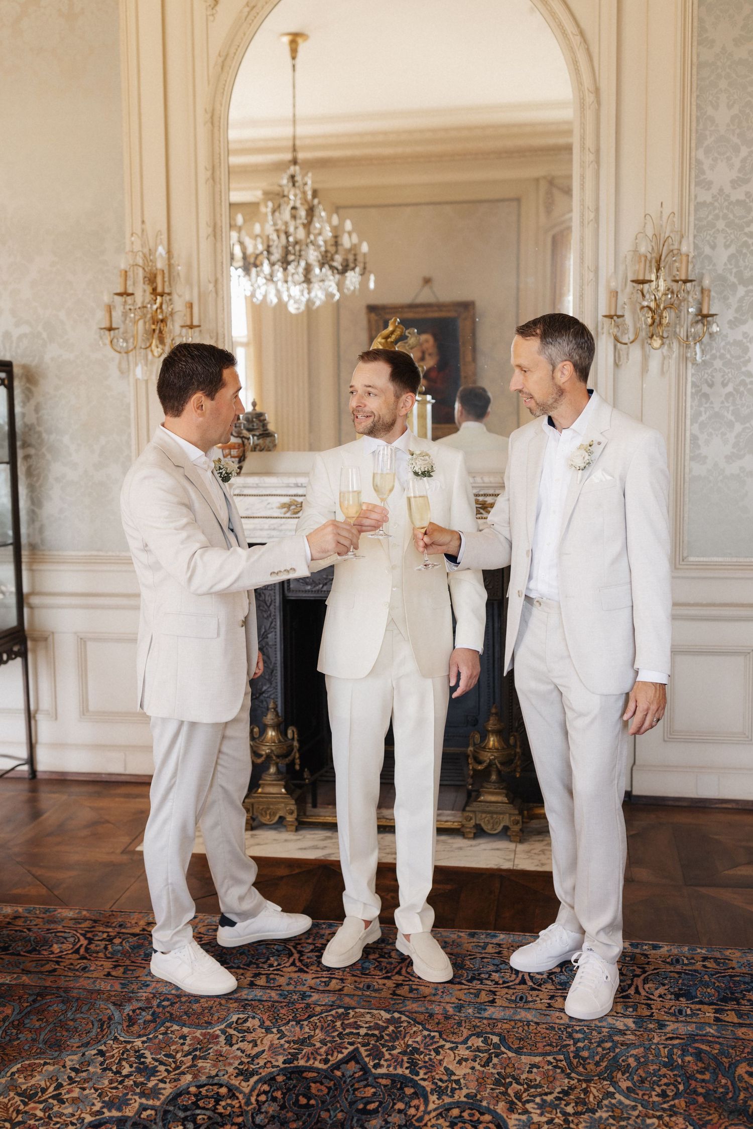 Groom and two groomsmen in white suits toasting champagne in grand ornate wedding venue.