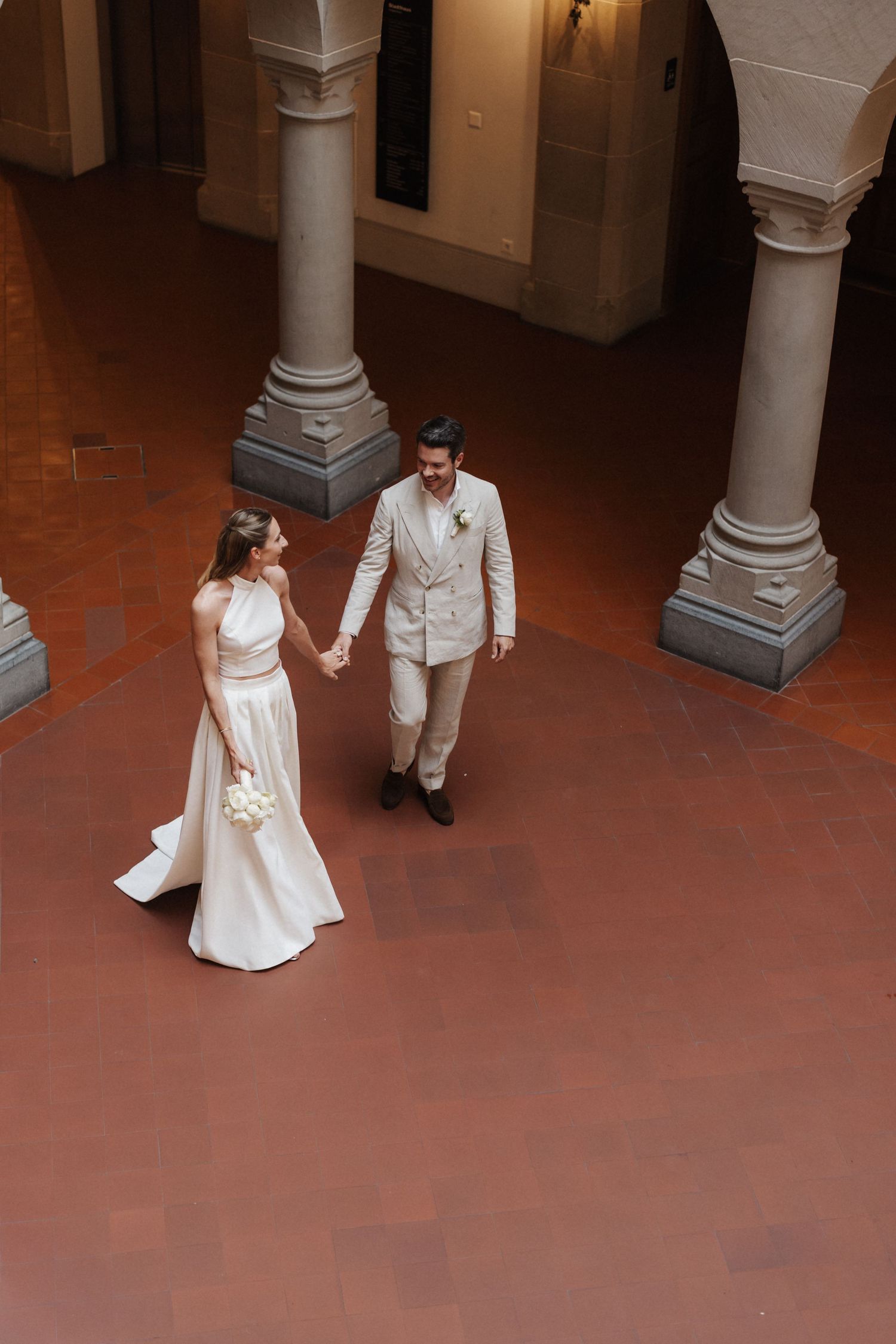 Bride and groom hold hands walking through elegant columned courtyard on their wedding day.