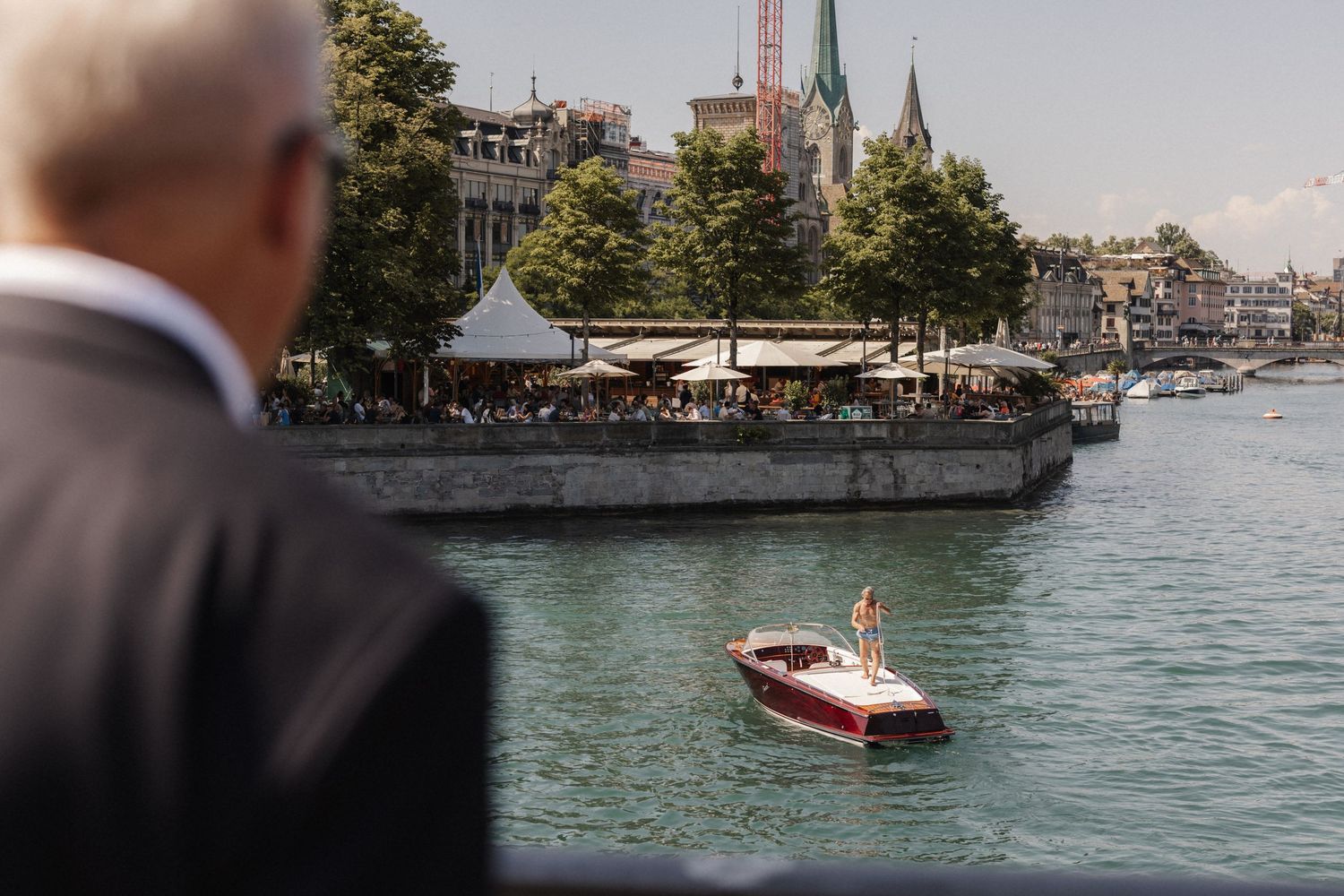 Wedding guests watch classic red motorboat cruise along scenic Zurich waterfront.