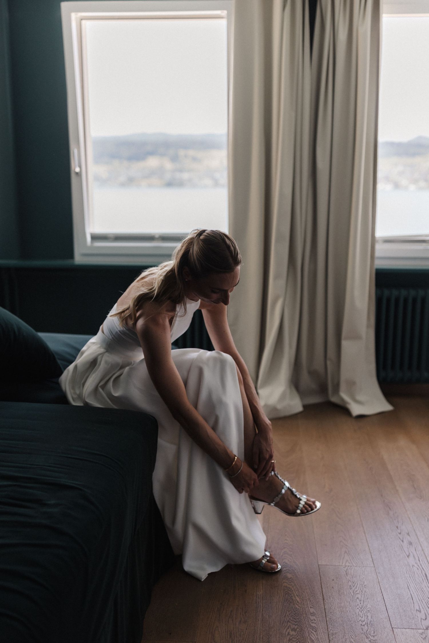 A bride in a white gown sits on a dark sofa by a window, fastening her silver heeled sandals.