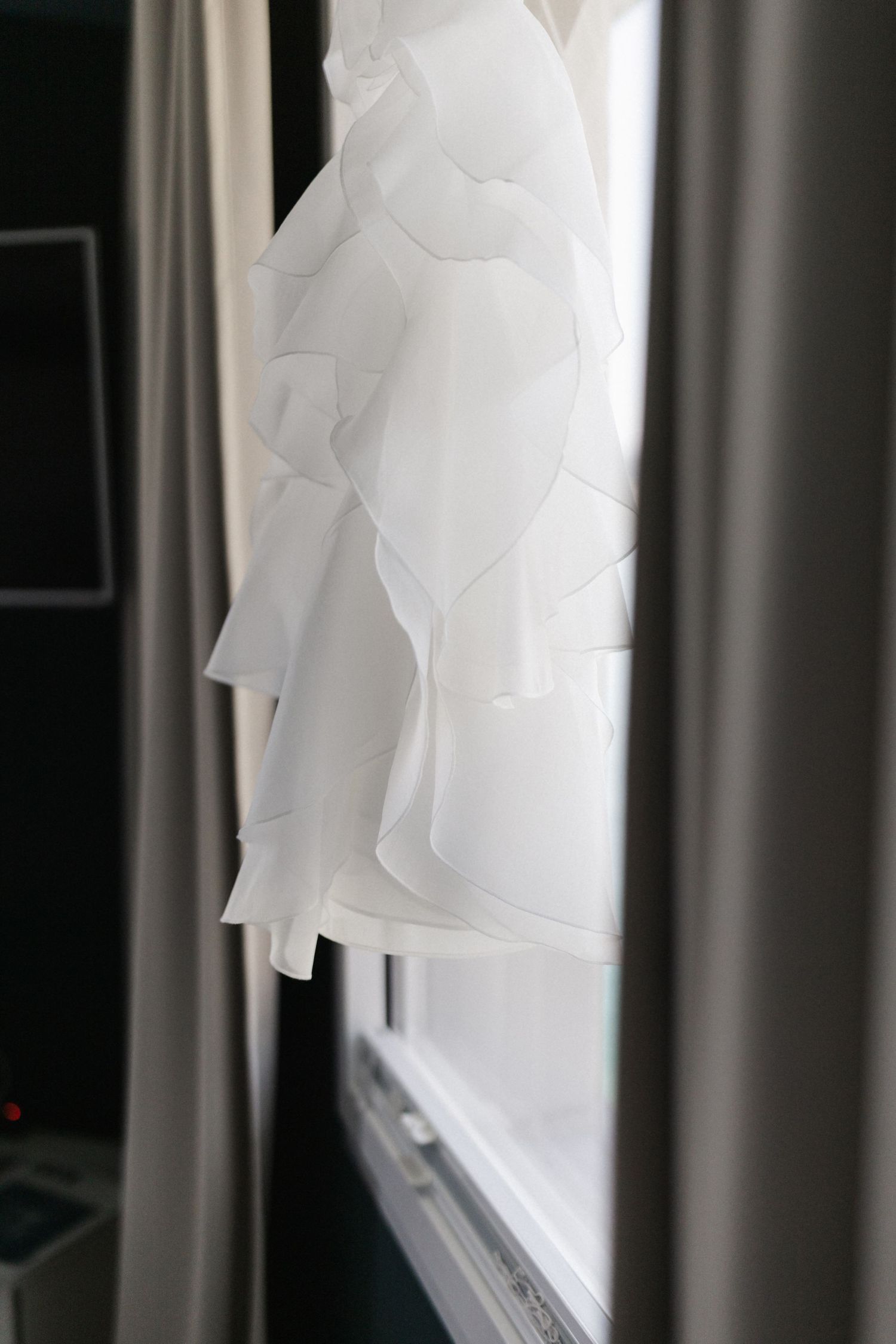 White ruffled wedding dress hanging against window in dark room.