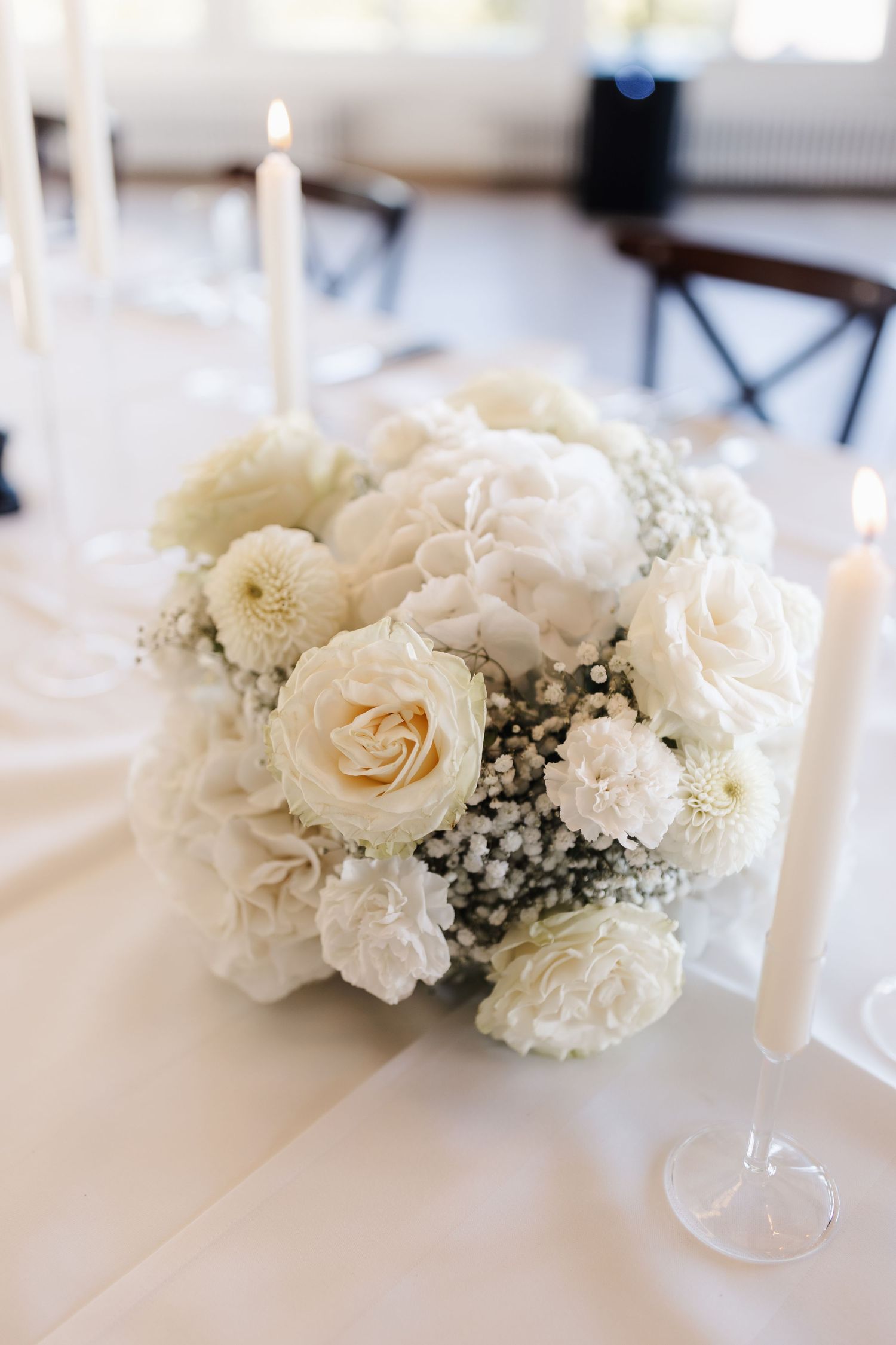 White wedding centerpiece of roses, carnations and baby's breath flanked by lit taper candles on white tablecloth.