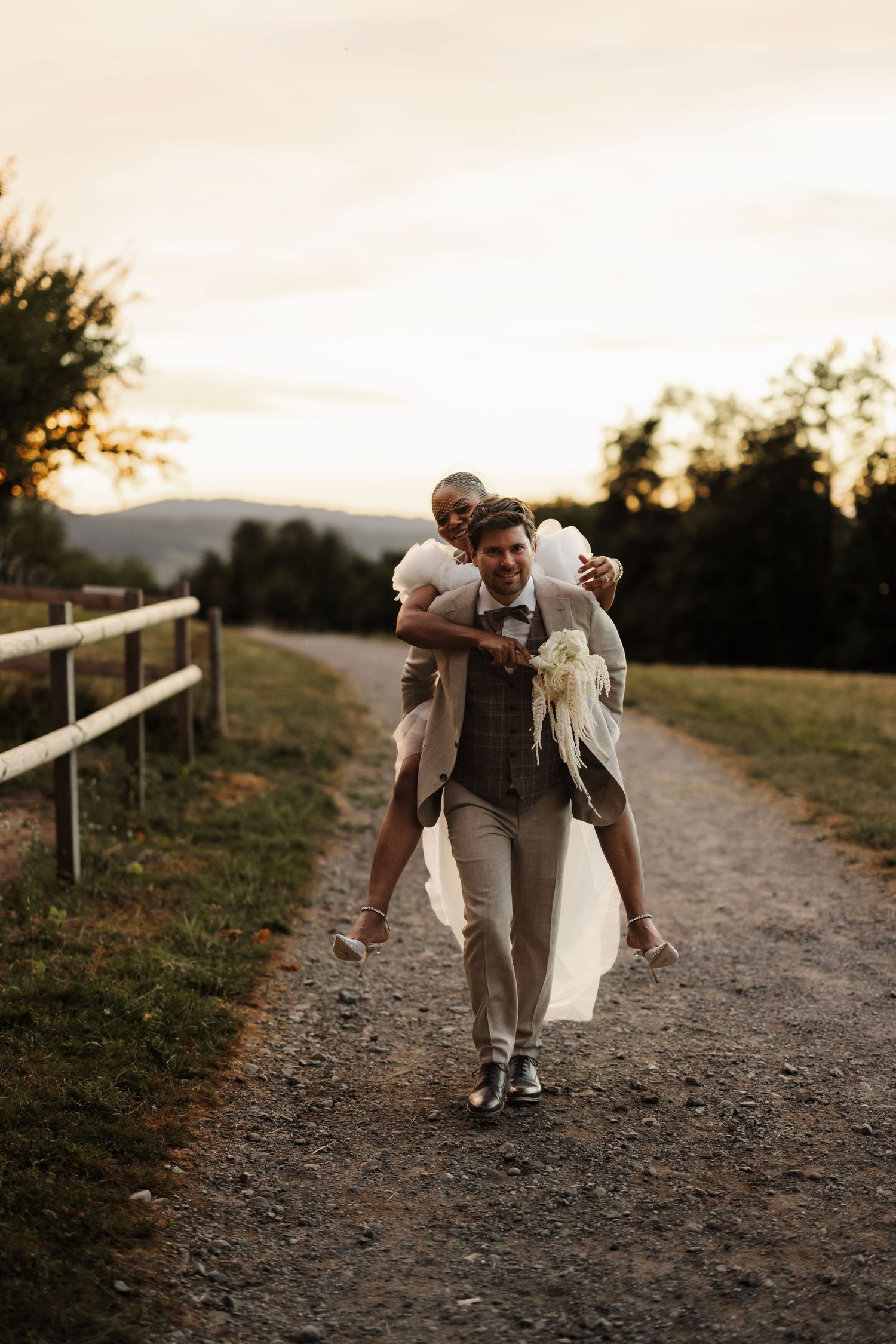 Groom giving bride a piggyback ride along a rustic gravel path at sunset, both laughing joyfully.