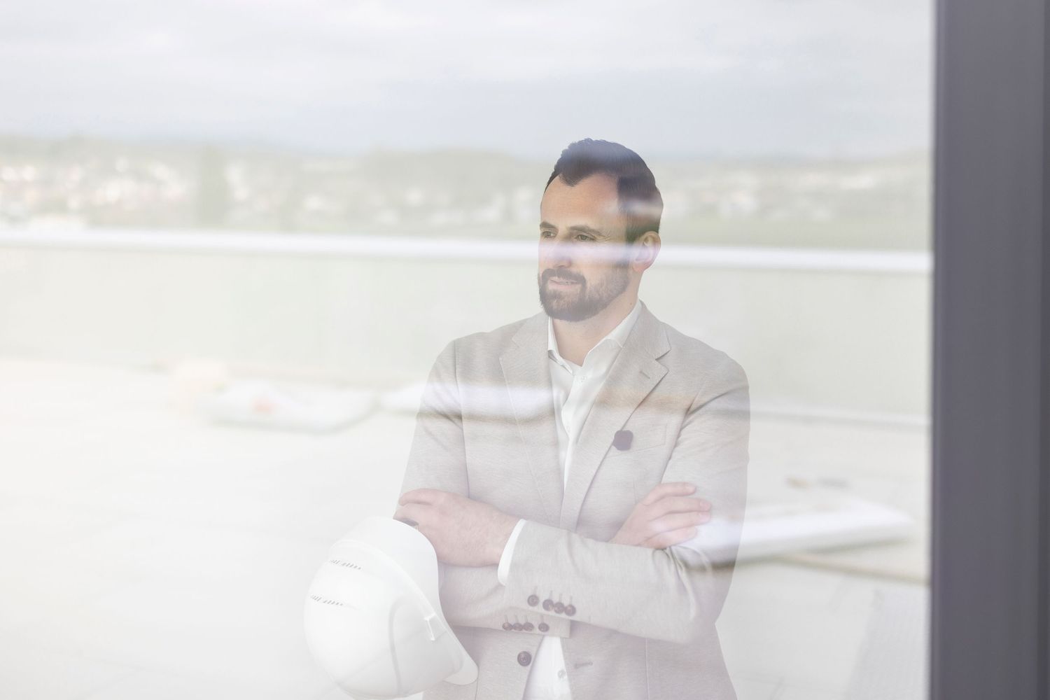 Confident businessman in beige suit holding a hard hat with arms crossed, viewed through a glass window.