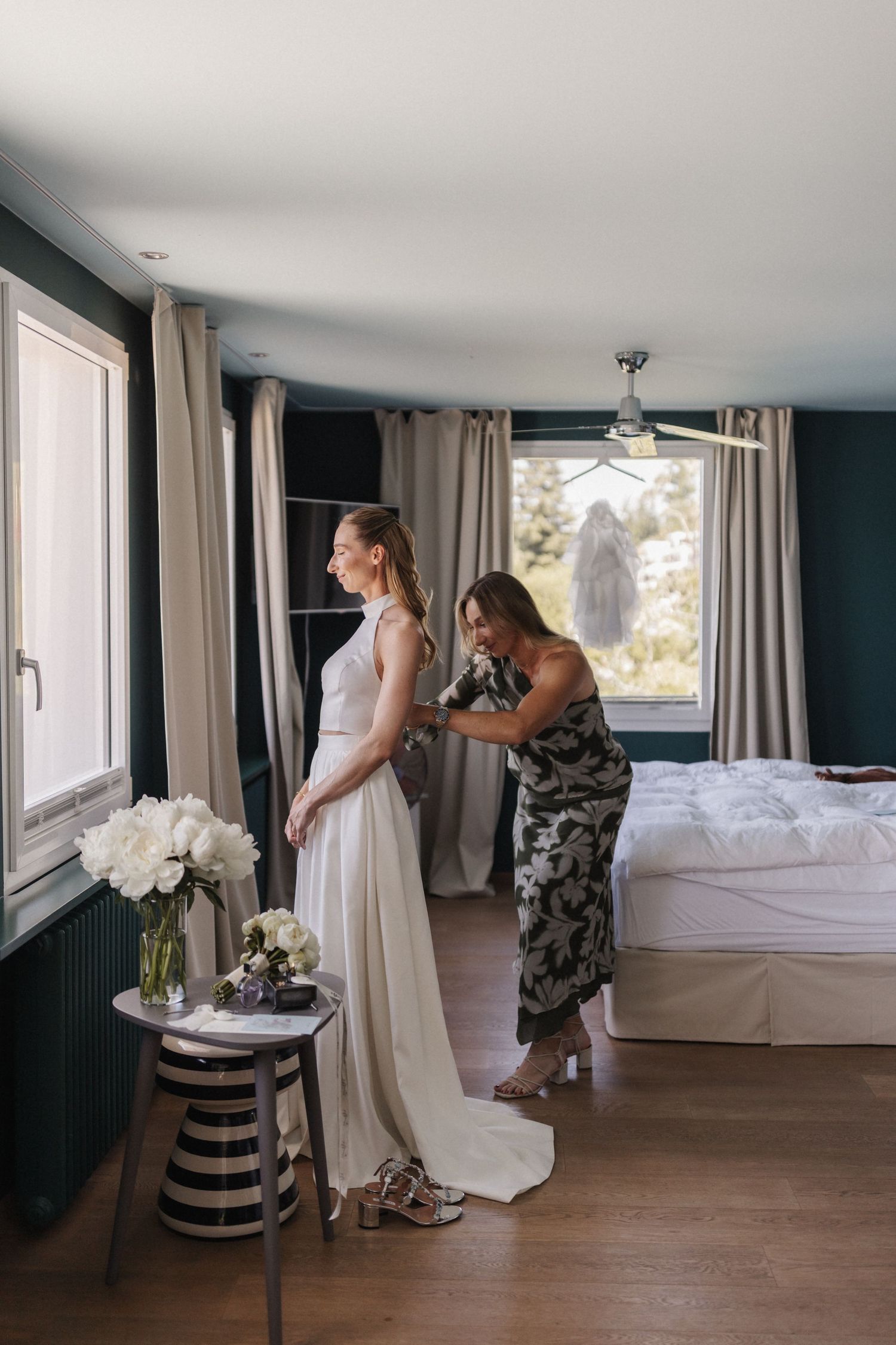 Bridesmaid in floral dress helping bride in white gown get dressed in a stylish bedroom.