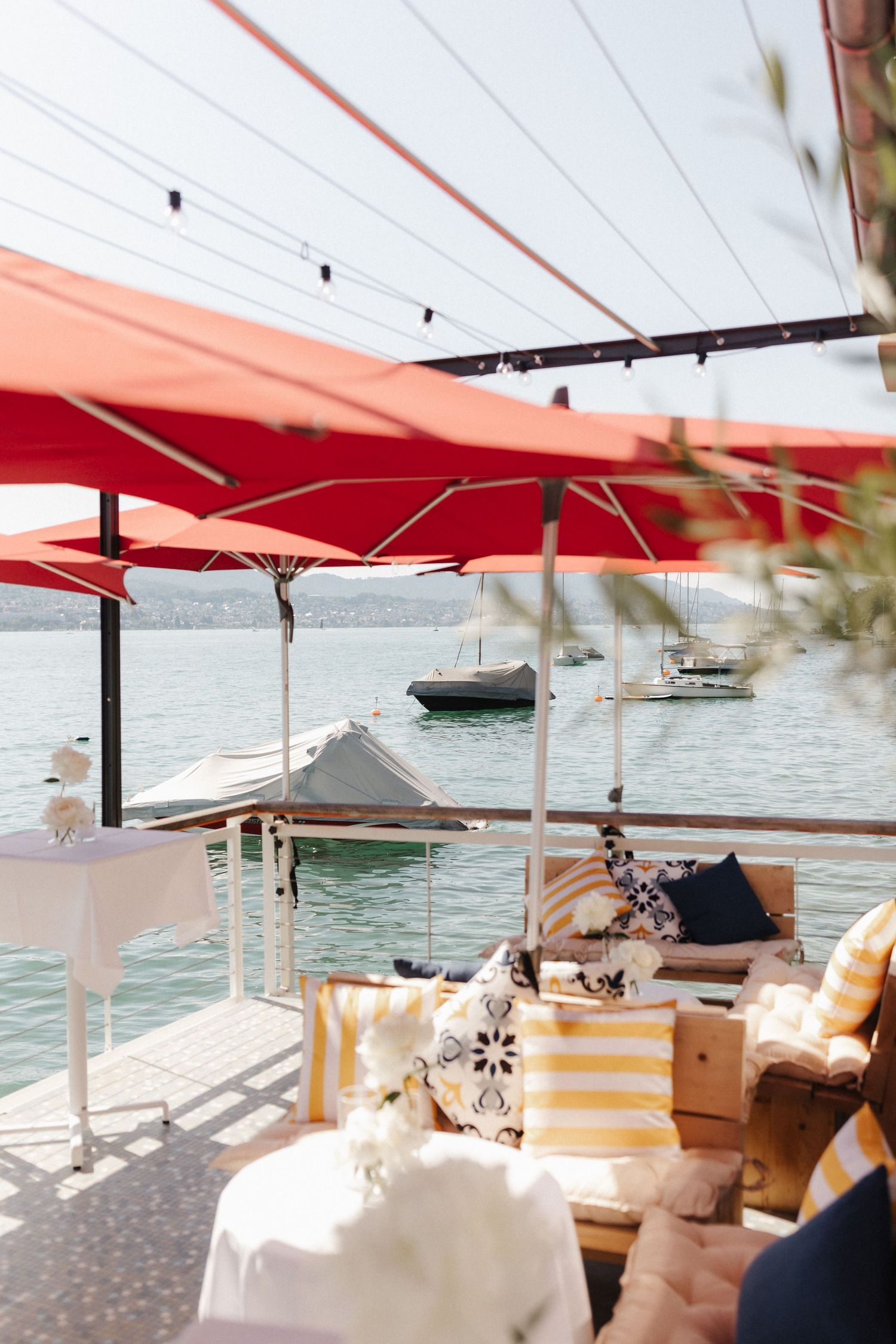 Elegant waterfront terrace with red umbrellas, striped cushions, and boats on calm blue water.