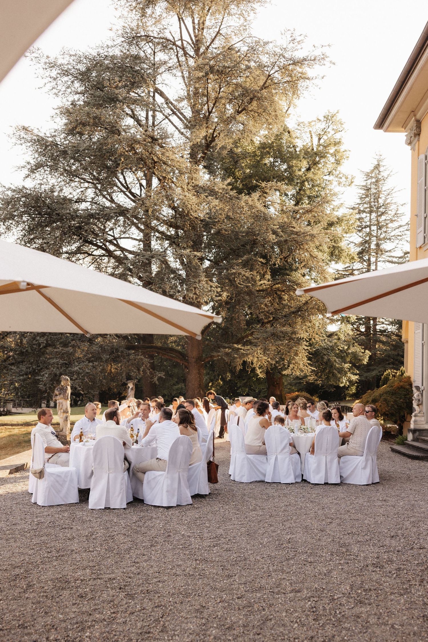 Outdoor wedding reception with white-dressed guests dining under cream parasols beside a classic European villa.