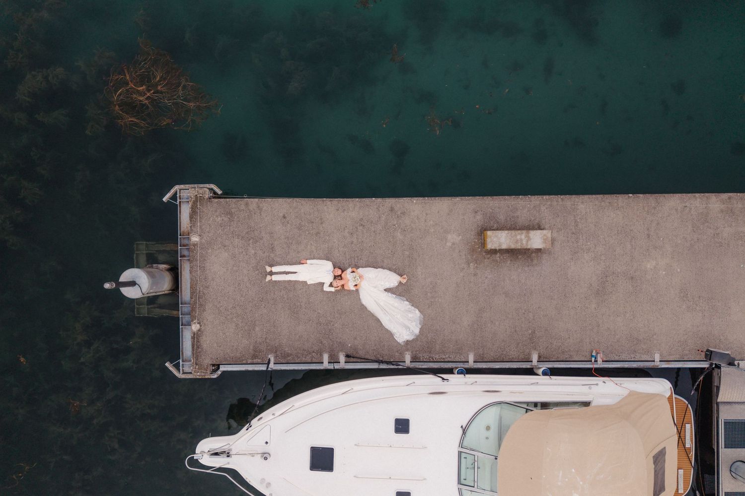 Top-down aerial shot of bride in white dress lying on a pier next to a moored boat.